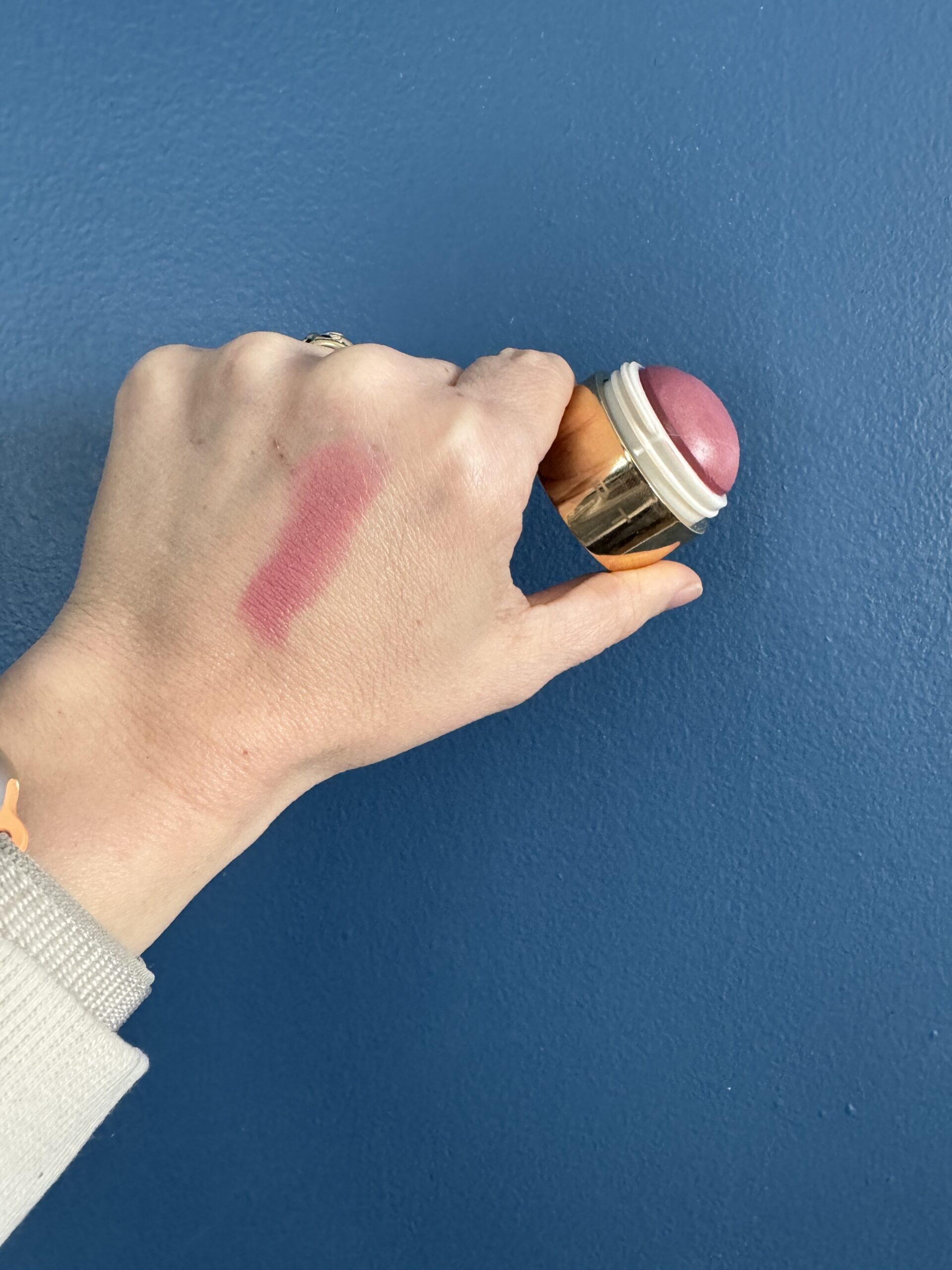 A hand holding a cosmetic product against a blue background with a swatch of pink makeup on the skin.