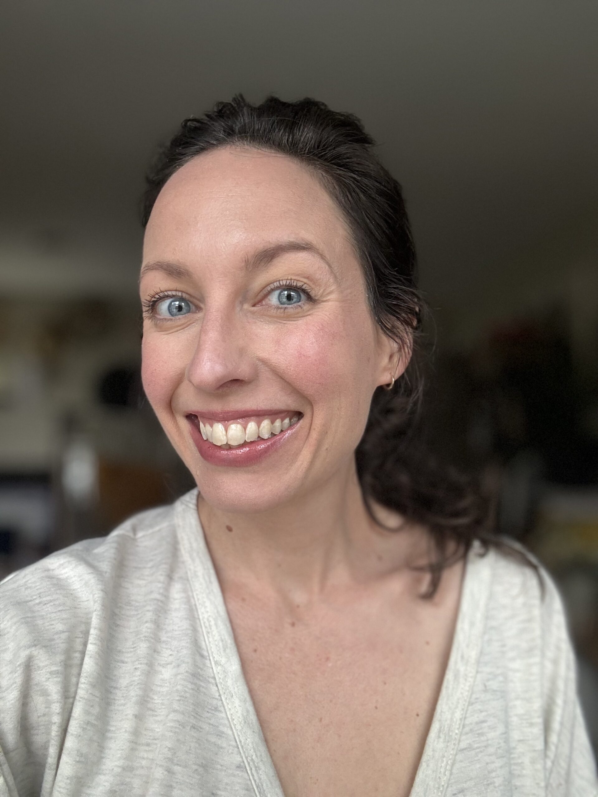 A woman with a joyful expression, wearing a light-colored top, poses for a selfie indoors.