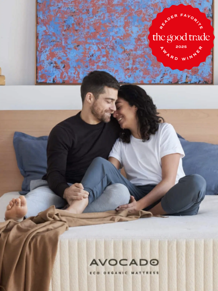 A couple sits on an Avocado Eco Organic Mattress, smiling at each other. A red badge in the corner reads "The Good Trade 2025 Reader Favorite Award Winner.