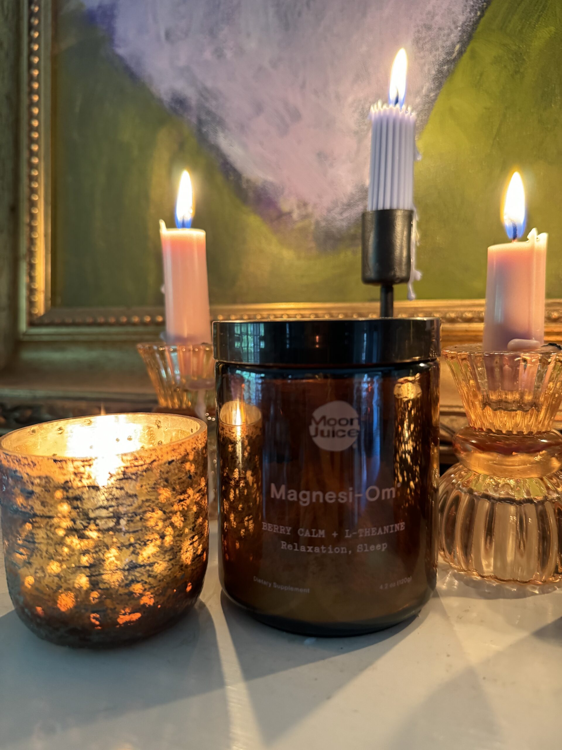 A candlelit setting with a jar labeled "moon juice magnesi-om" and three lit candles, two in glass holders and one mounted on a metal stand, against a green painting background.