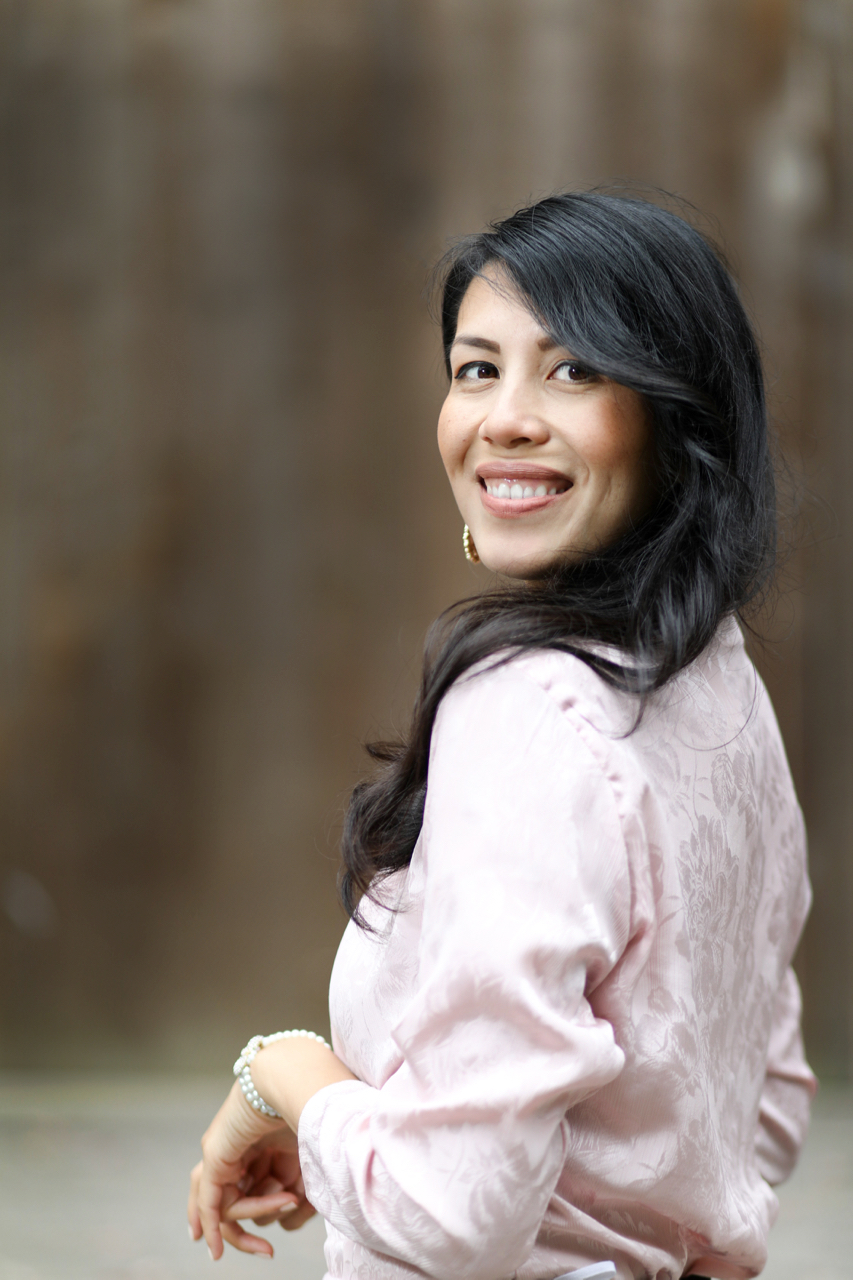 A smiling woman with dark hair wearing a pink blouse and pearl bracelet, looking over her shoulder against a blurred background.