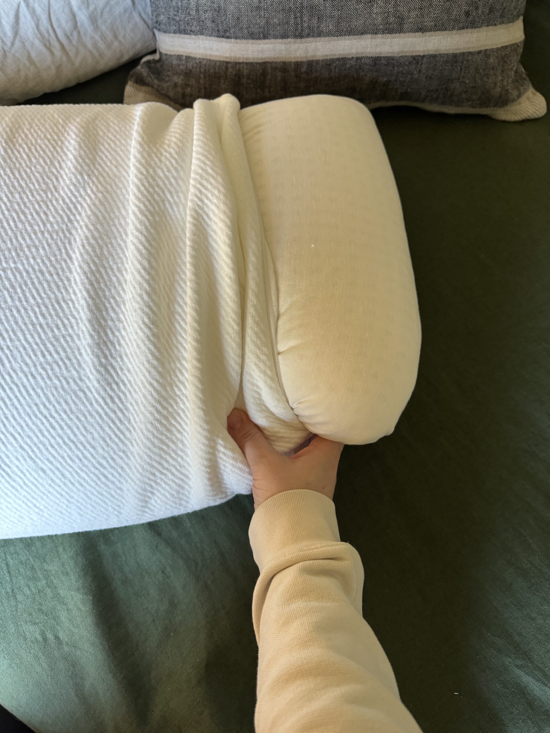 A person is tucking in a white blanket on a bed or couch with various cushions.