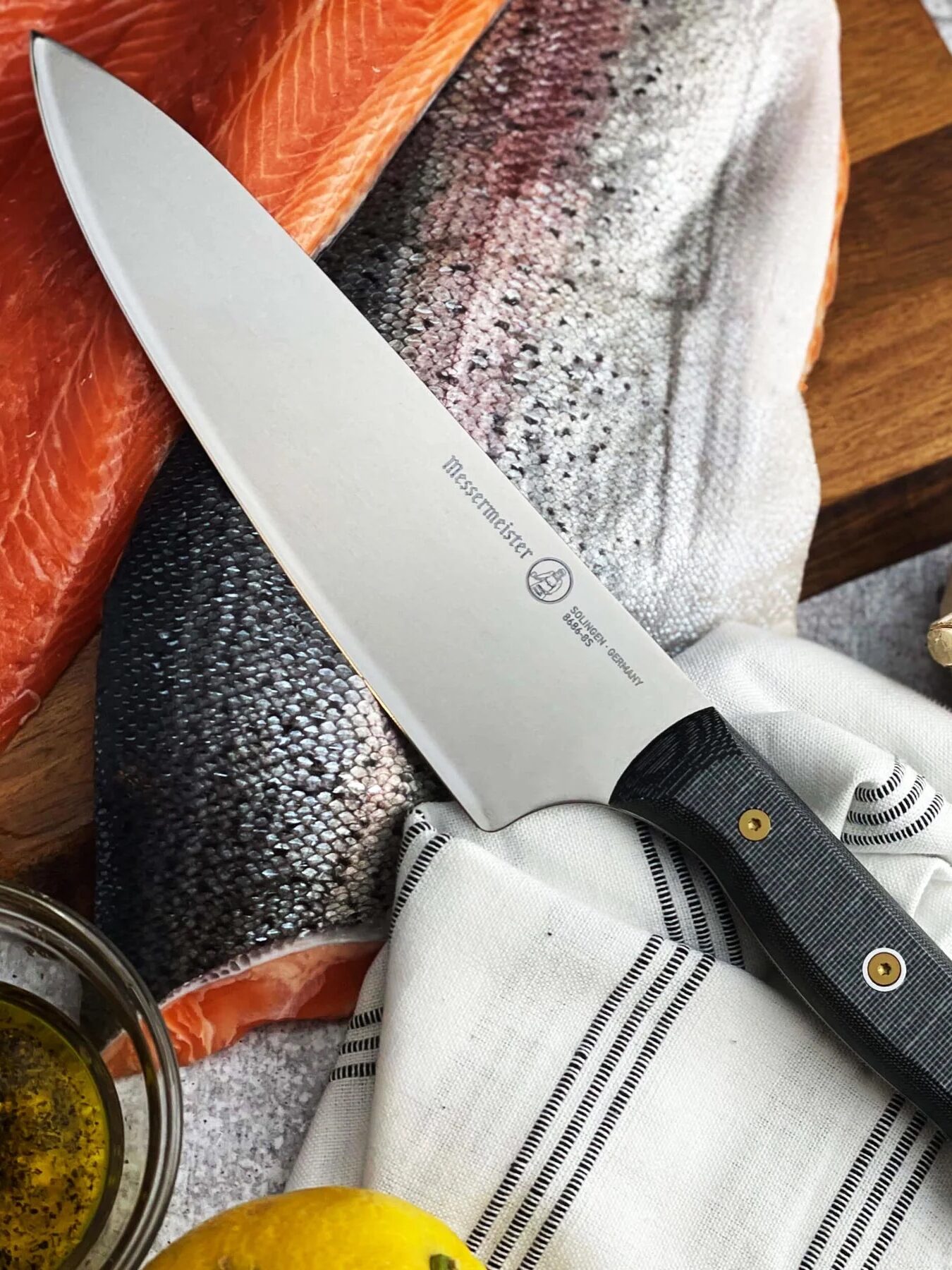 A chef's knife rests on a white cloth next to raw salmon, garlic, lemon, and herbs, all arranged on a gray textured surface.