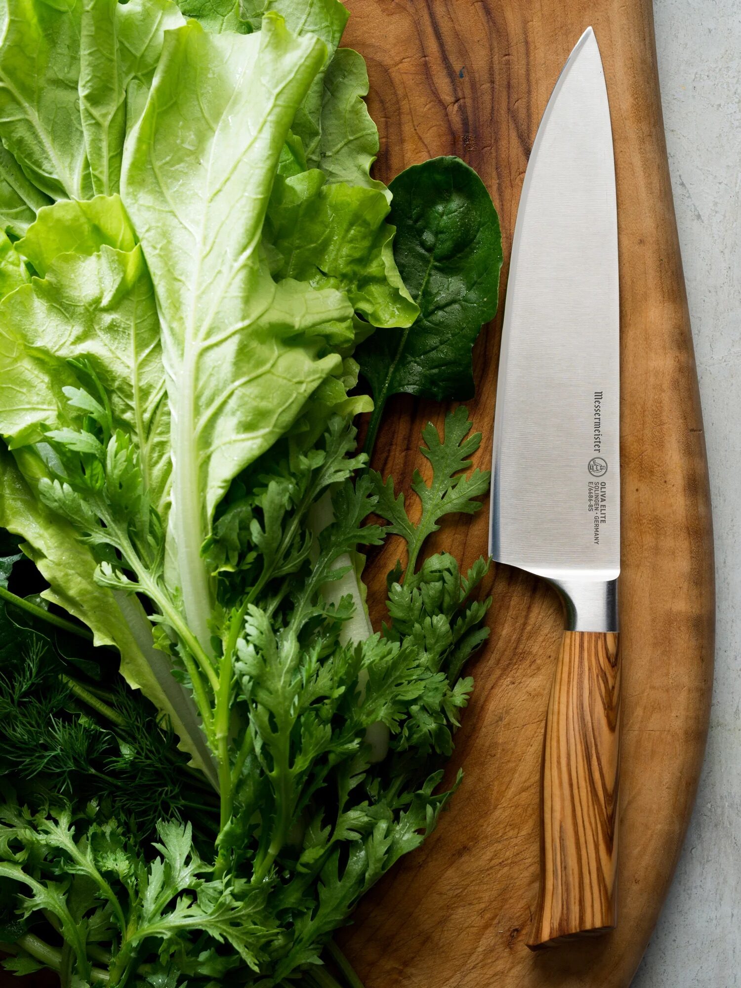 A chef's knife on a wooden cutting board surrounded by fresh green lettuce and herbs.