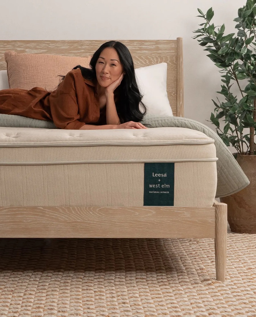 A person leisurely lounges on a bed with a nontoxic Leesa West Elm mattress, surrounded by plush pillows and a vibrant plant.
