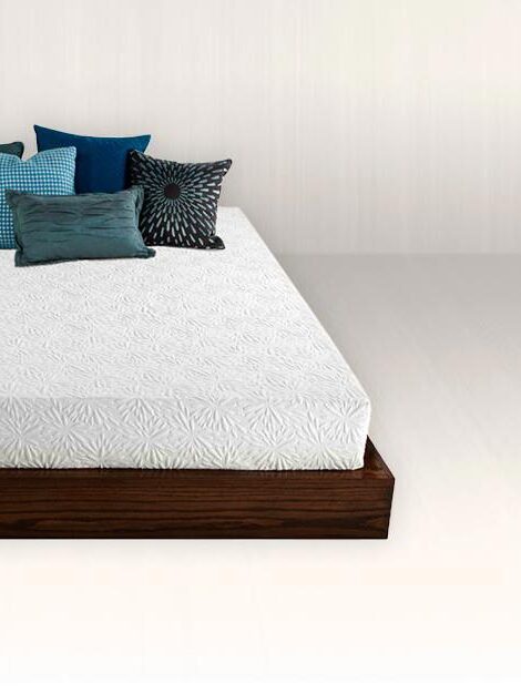 A modern low wooden bed frame with a white mattress covered by several plush pillows in various shades of blue and patterns, positioned in a room with a plain white floor and wall.