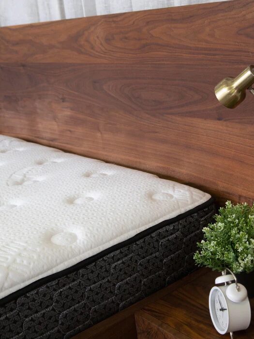 A modern bedroom highlighting a walnut headboard, white quilted mattress, black textured bed base, brass reading lamp, and a bedside table with a white alarm clock and a potted plant.
