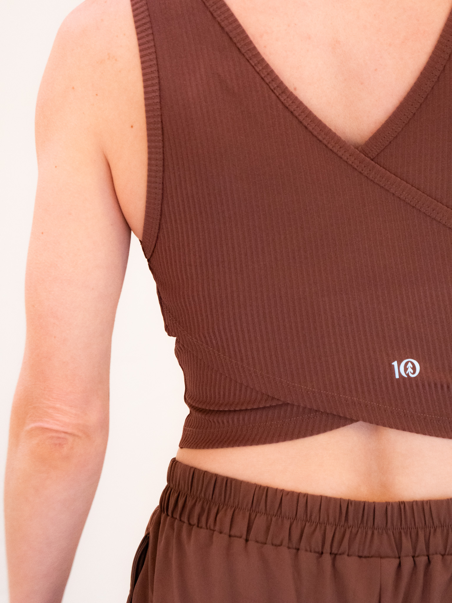 Close-up of a woman wearing a brown sports bra, displaying the logo on the lower left side, against a light background.