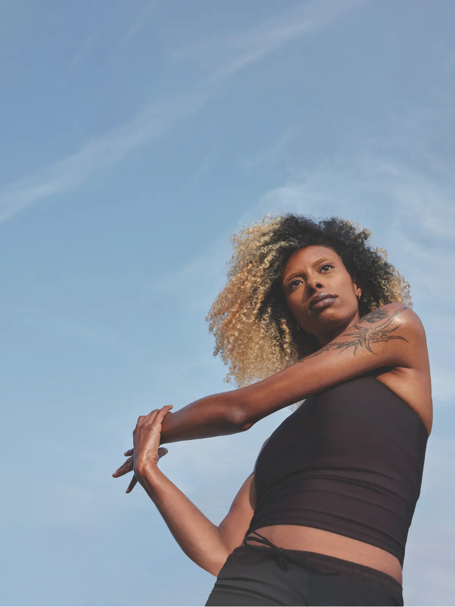 A young woman with curly blonde hair stands against a blue sky, looking contemplative, wearing a black top and showing a tattoo on her arm.