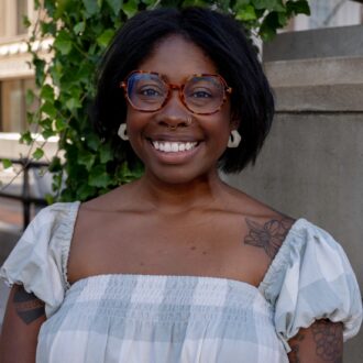 A person with short black hair and glasses stands outdoors next to some green ivy, wearing a light-colored off-the-shoulder top and smiling.