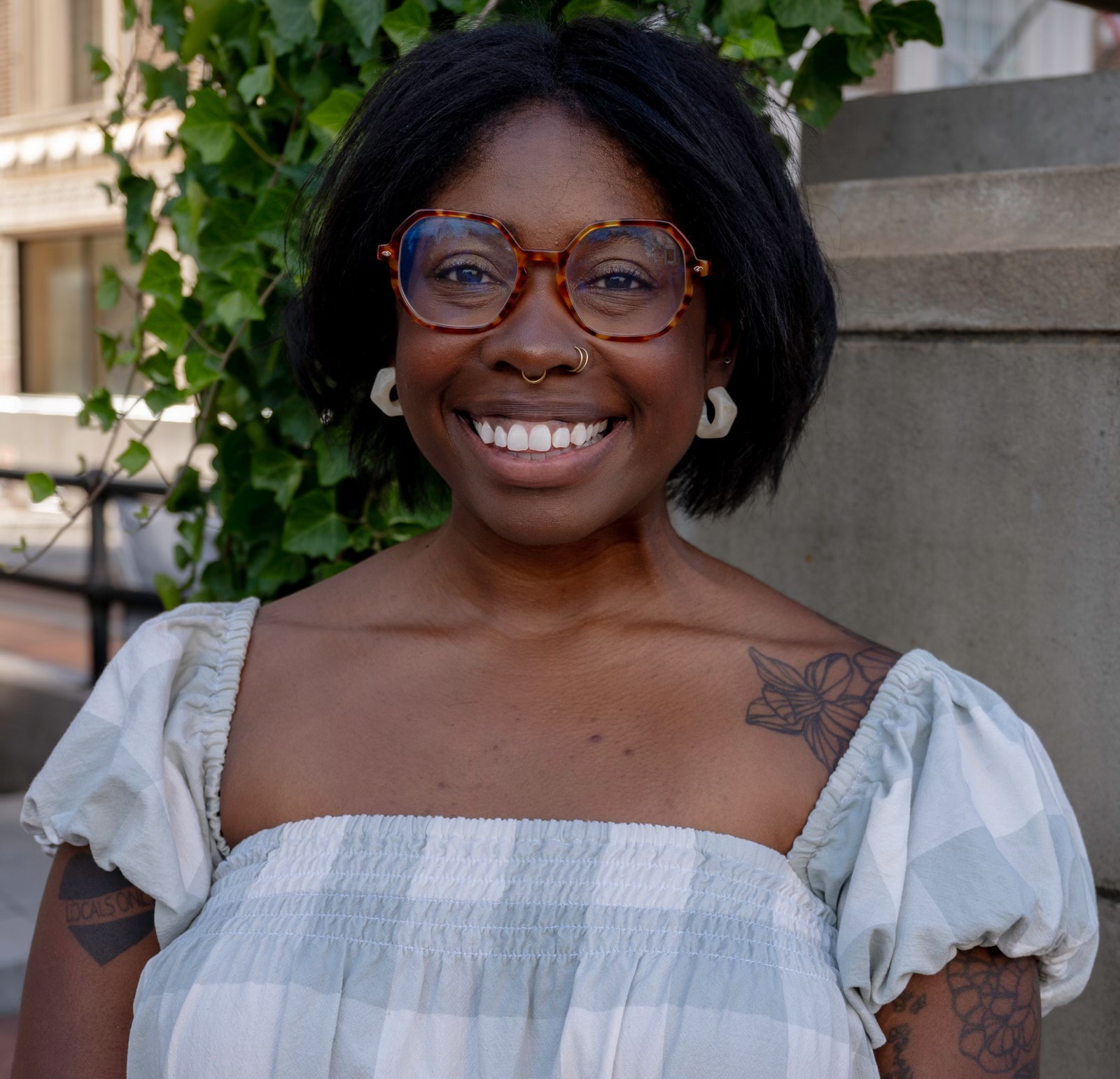 A person with short black hair and glasses stands outdoors next to some green ivy, wearing a light-colored off-the-shoulder top and smiling.