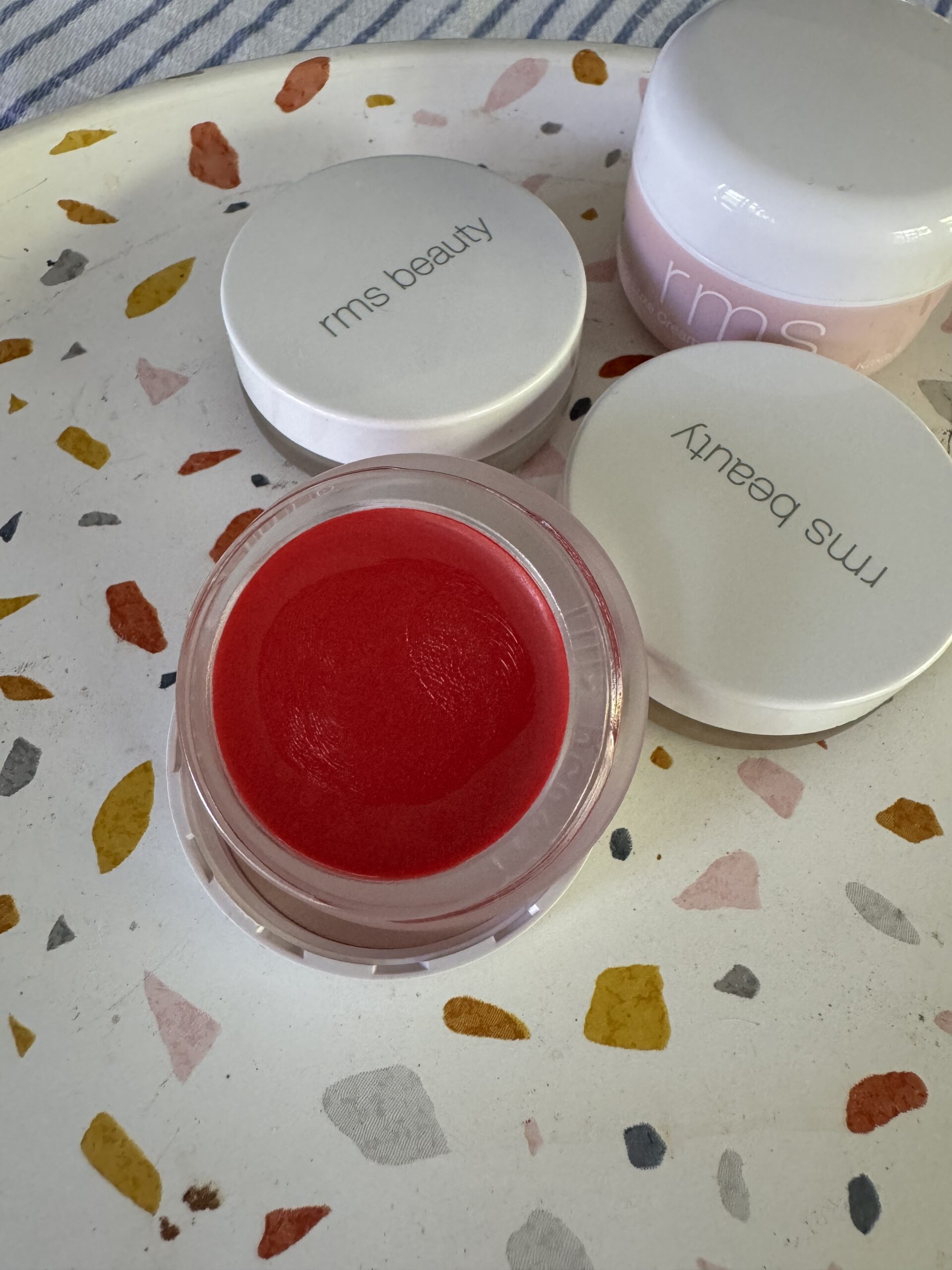 A circular container of red lip balm labeled "rms beauty" is open on a speckled surface, with two closed containers, also labeled "rms beauty," nearby.