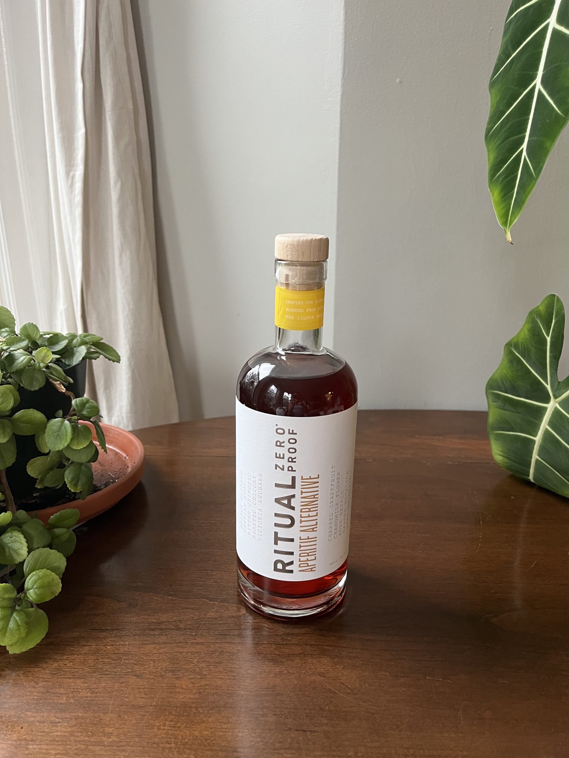 A glass bottle of Ritual Zero Proof whiskey alternative stands on a wooden surface. The label is white with black text. Green plants are in the background.