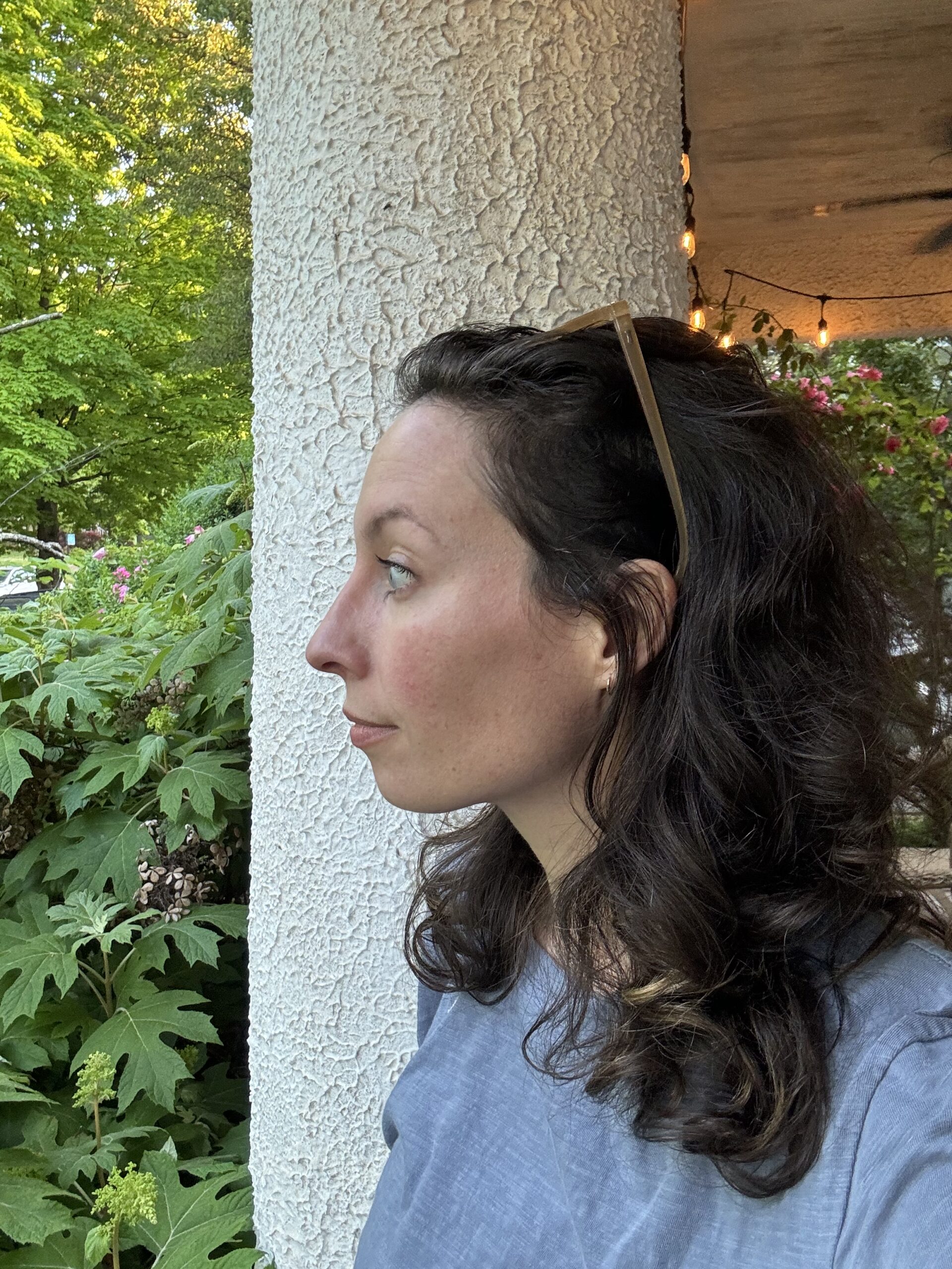 Profile view of a woman with curly hair, using a hair clip, standing next to a textured column with green foliage in the background.