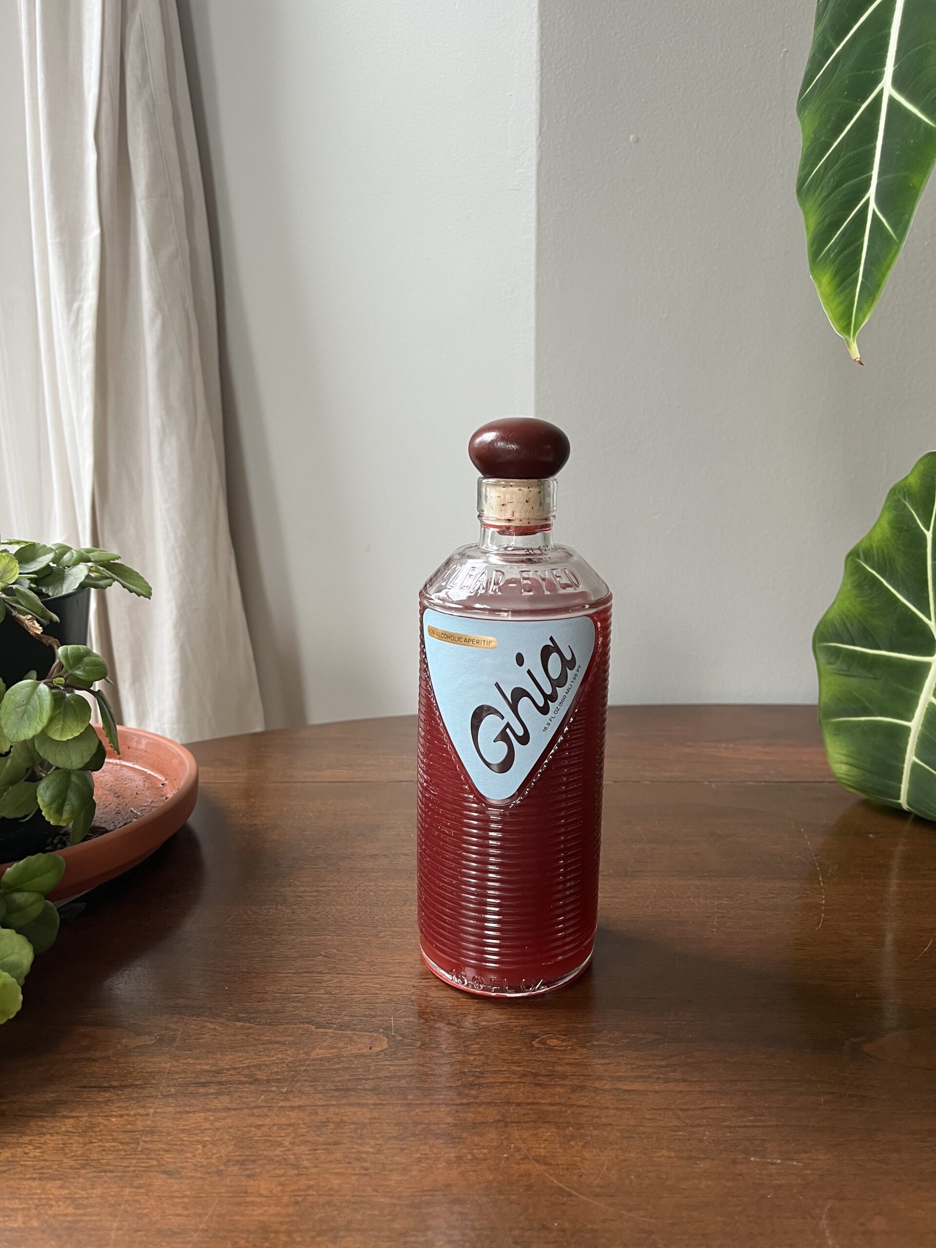 A bottle of Ghia non-alcoholic aperitif with a wooden cap sits on a wooden table. Green potted plants are placed around it.