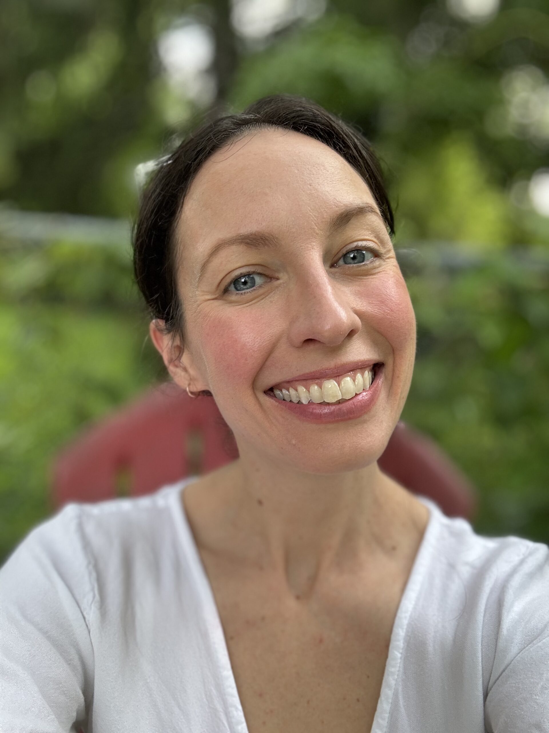 A person with light skin, blue eyes, and dark hair smiles at the camera, wearing a white top and sitting outdoors with green foliage in the background.