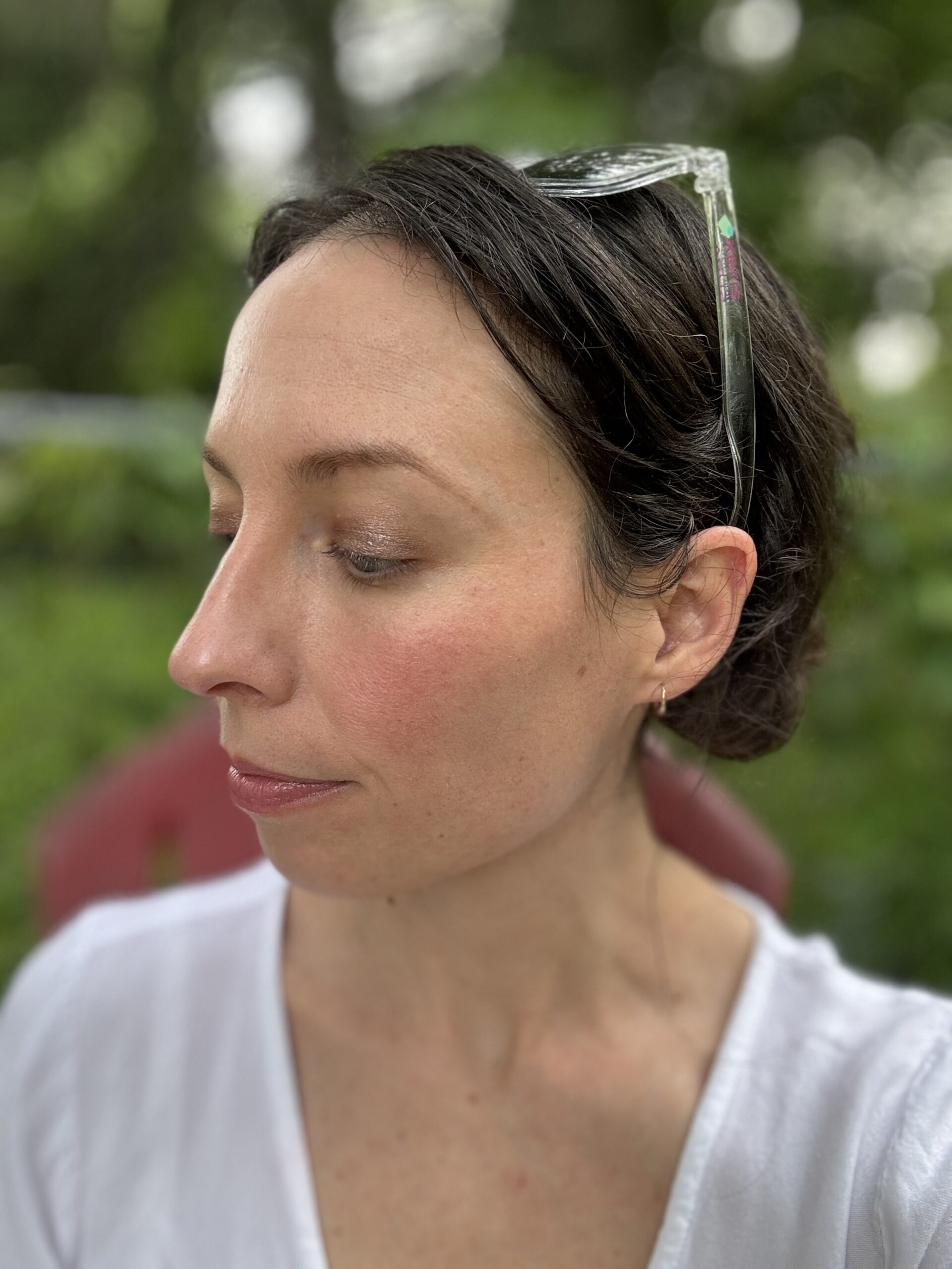 A woman with brown hair looks downward while seated outdoors. She is wearing a white shirt and has sunglasses resting on her head. The background is blurred greenery.