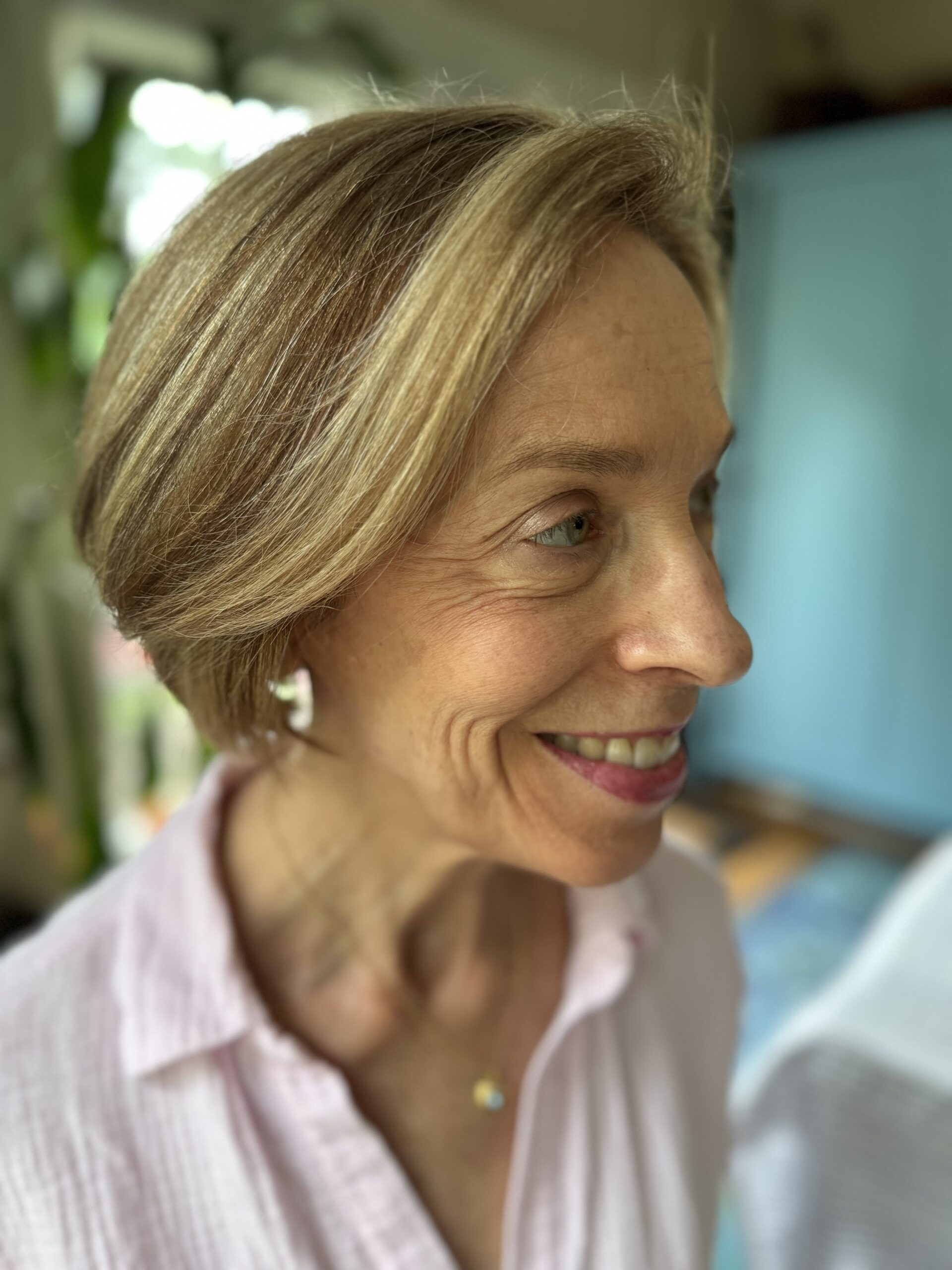 A woman with short blond hair and wearing a pink shirt smiles while looking to the side. She is indoors with a blue cabinet and greenery in the background.