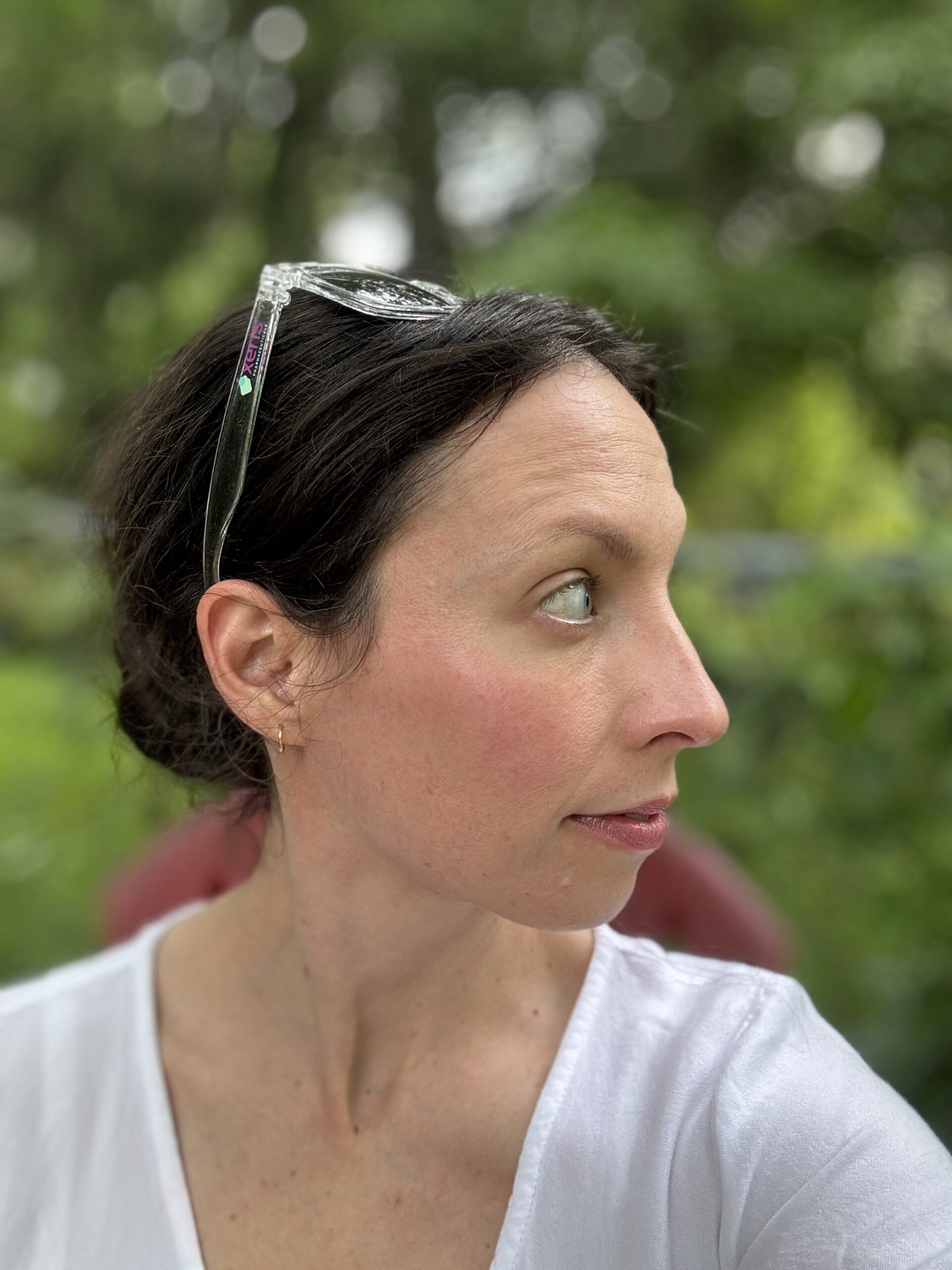 A person with dark hair, glasses resting on their head, and wearing a white shirt looks to the side with a neutral expression. The background is blurred greenery.
