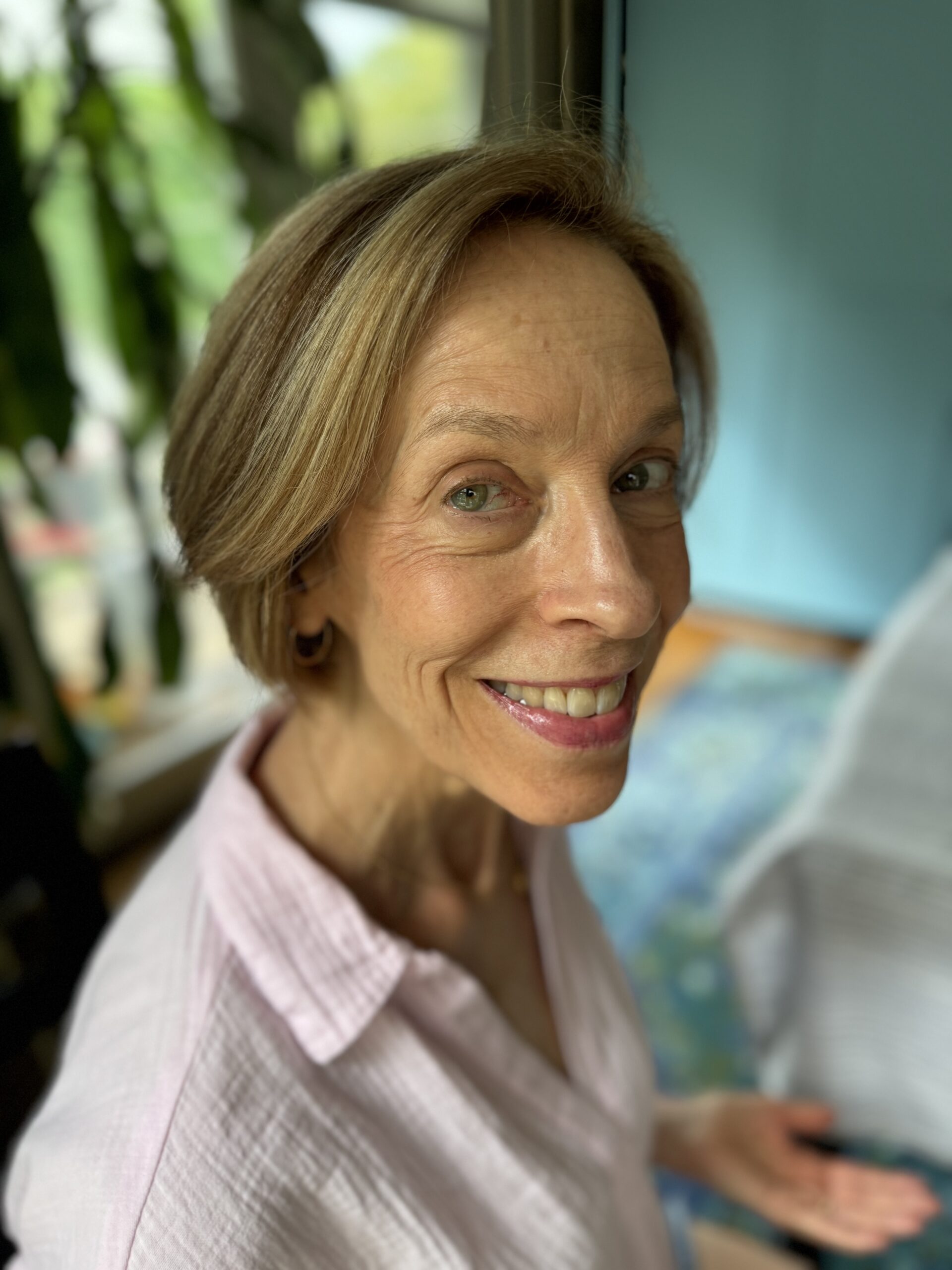 A woman with short, light brown hair wearing a light pink shirt is smiling while standing indoors near a window with green plants in the background.