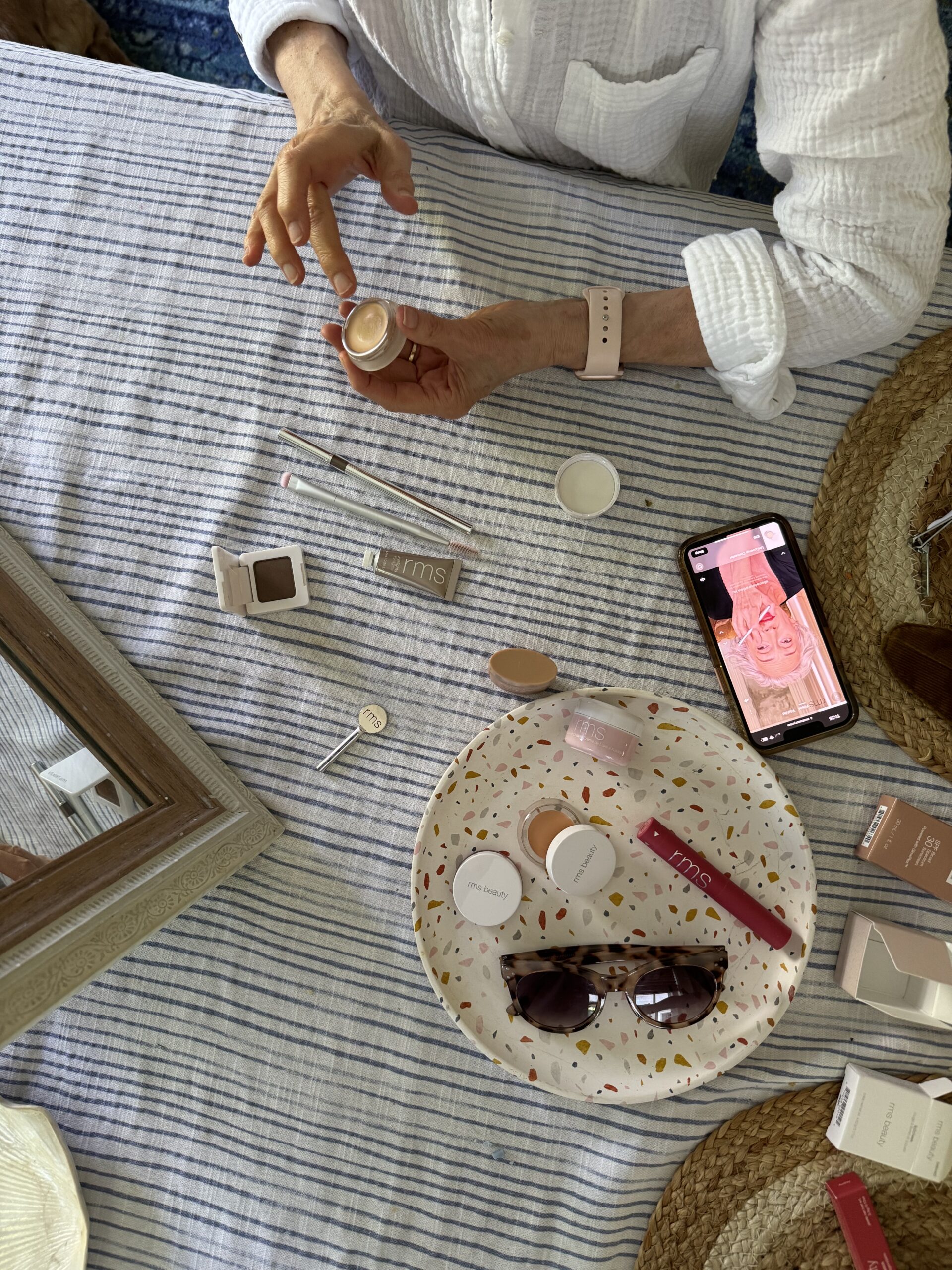 A person seated at a table applies a product from a small container. The table is covered with makeup items, a smartwatch, sunglasses, and a phone displaying an app.
