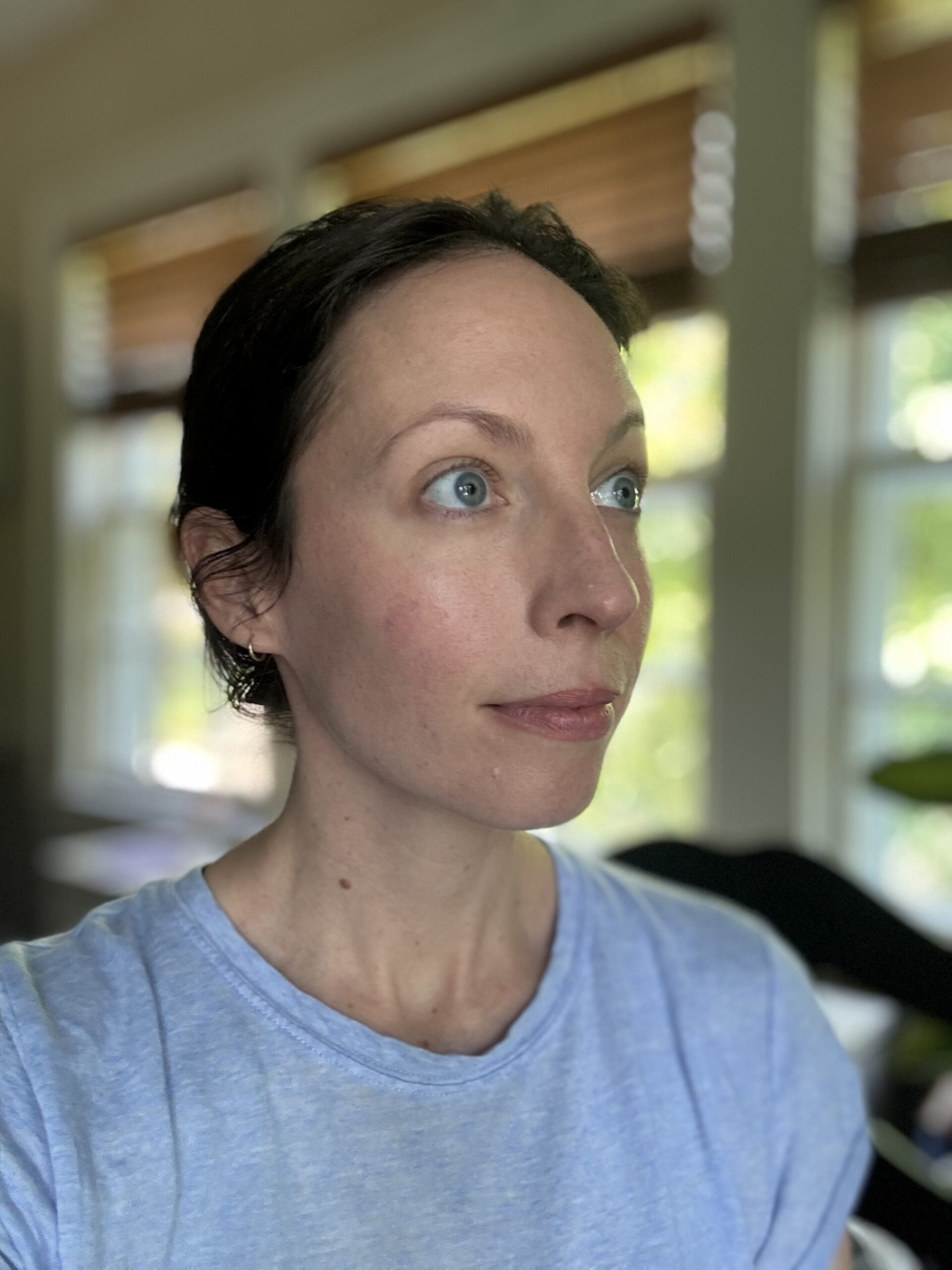 A woman with short dark hair and blue eyes is looking to the right. She is wearing a light blue shirt, with a background featuring windows and greenery.
