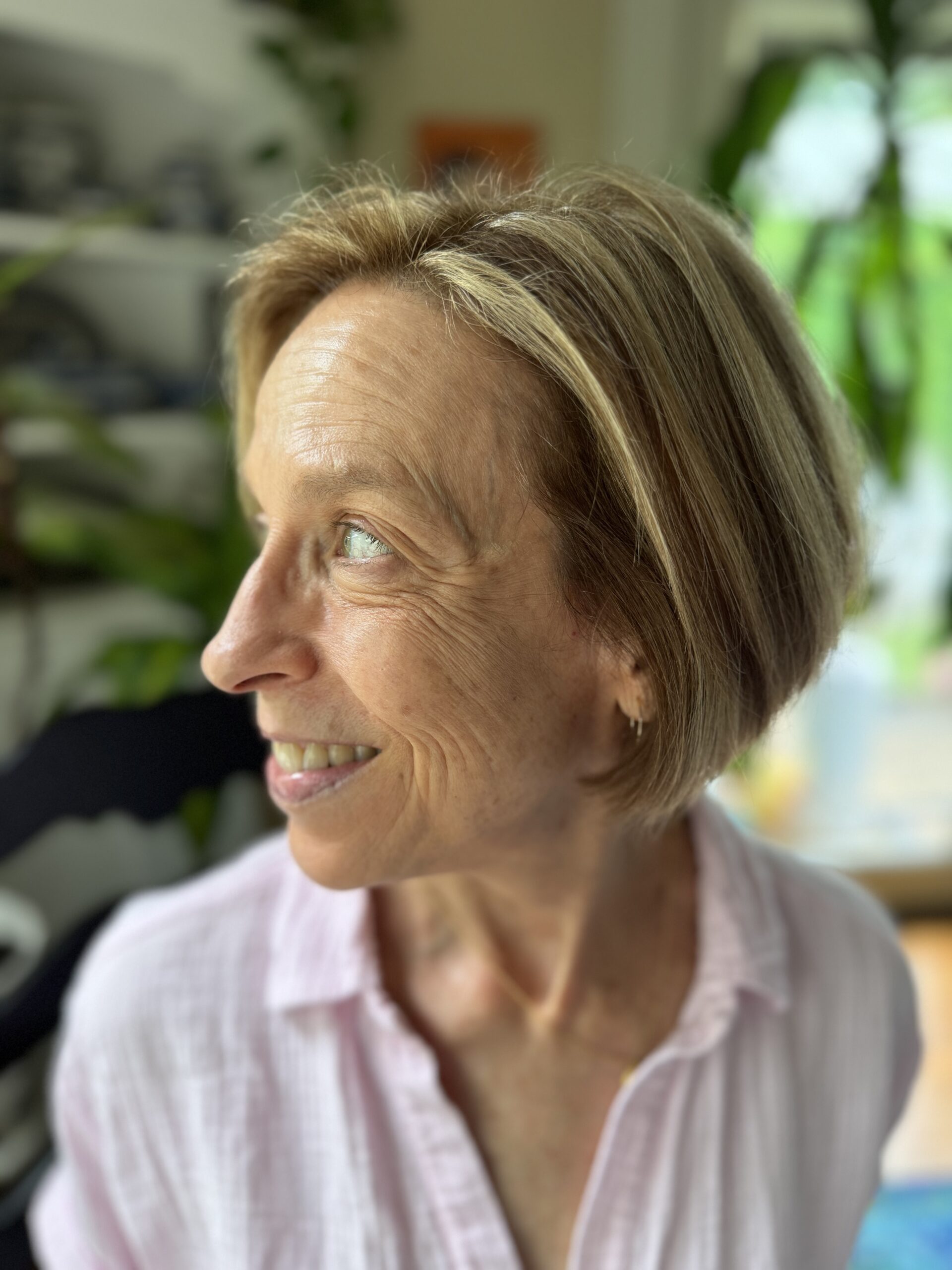 A woman with short blonde hair and a pink shirt looks to her left, smiling. She is indoors with green plants visible in the background.