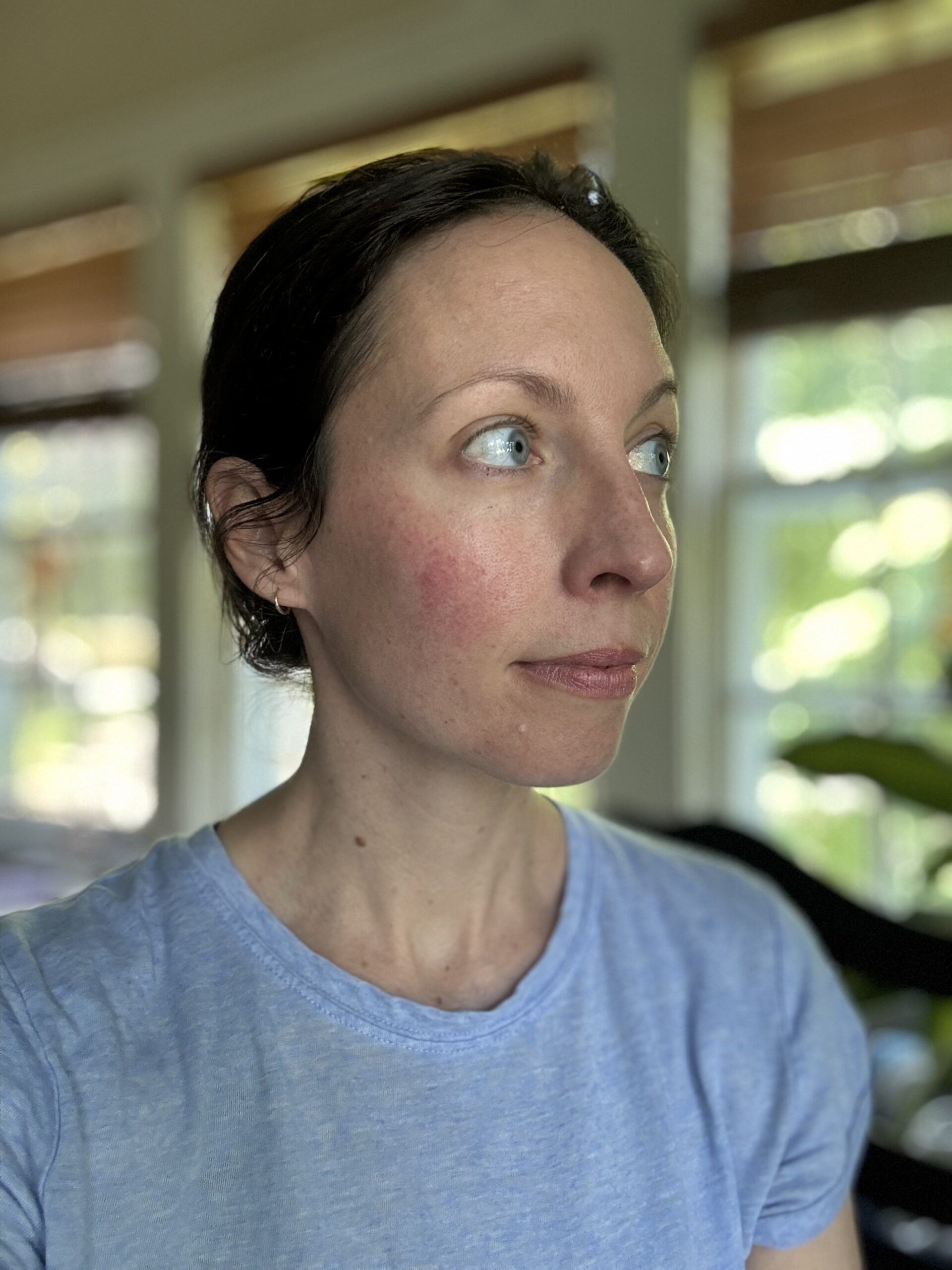 A person with short dark hair and fair skin, wearing a light blue shirt, looks to the side in a brightly lit room with large windows and greenery visible outside.