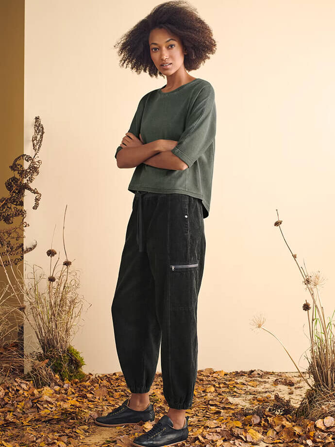 A person stands with arms crossed, wearing a green short-sleeve top and black pants, in front of a beige wall with dried plants and leaves scattered on the ground.