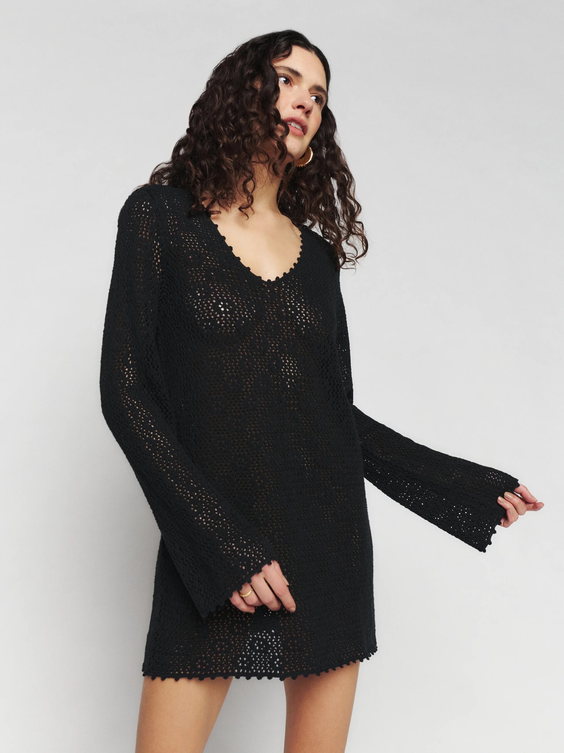 A person with curly hair wearing a black, long-sleeved, crochet dress poses against a plain background.