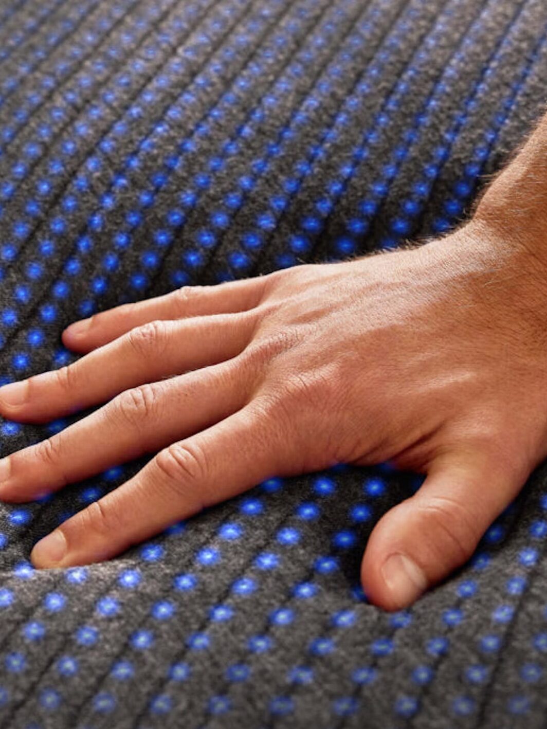 A hand pressing down on a grey mattress with blue dots, demonstrating the mattress's ability to conform to pressure.
