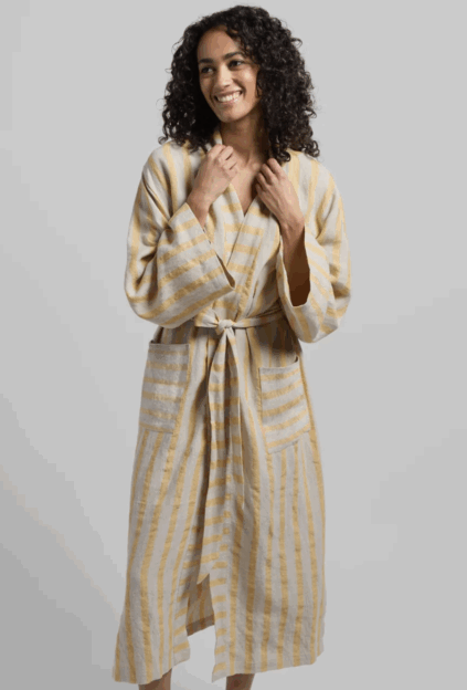 A person wearing a long, striped bathrobe with a tie belt stands against a plain background, smiling and looking to the side.