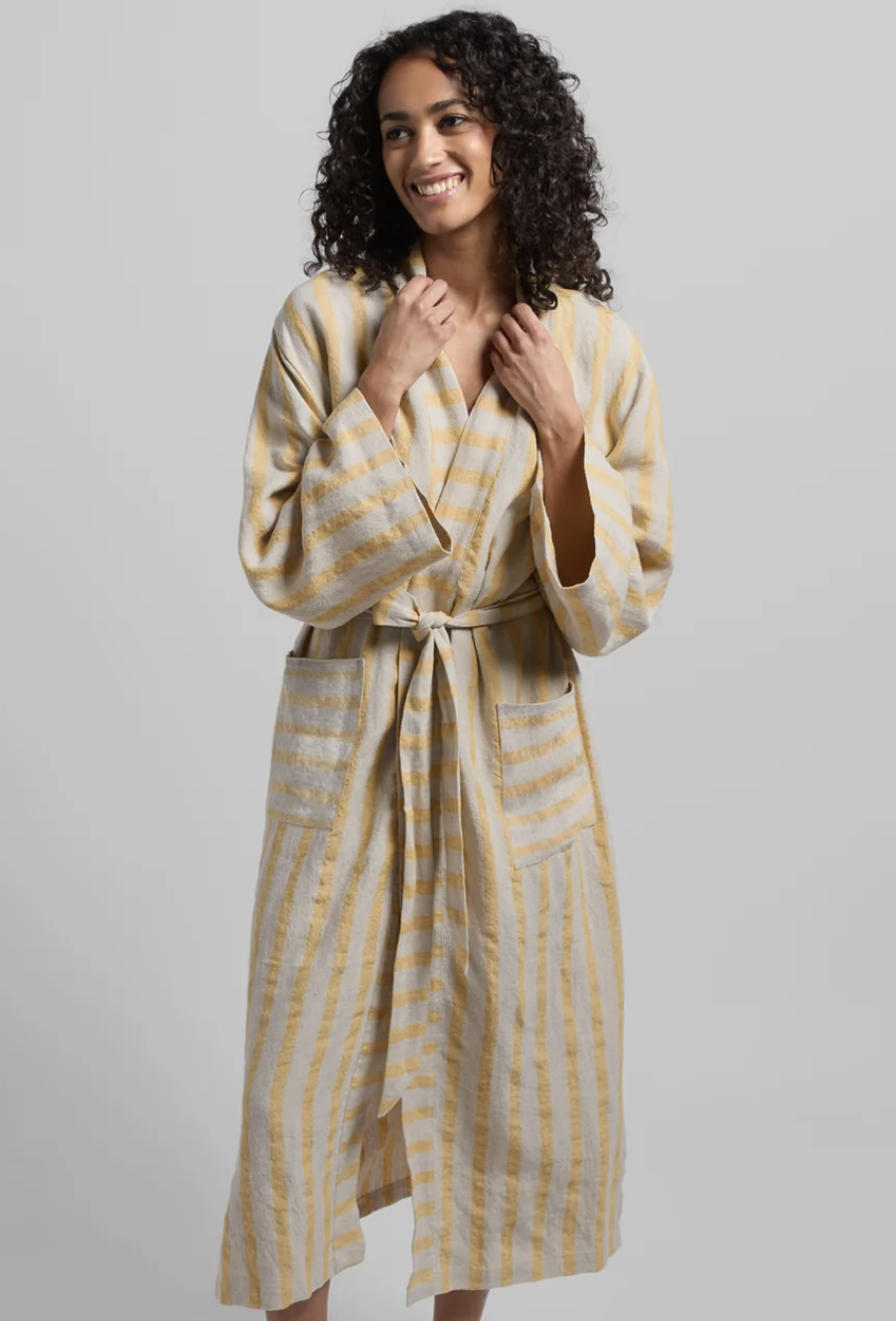 A person wearing a long, striped bathrobe with a tie belt stands against a plain background, smiling and looking to the side.