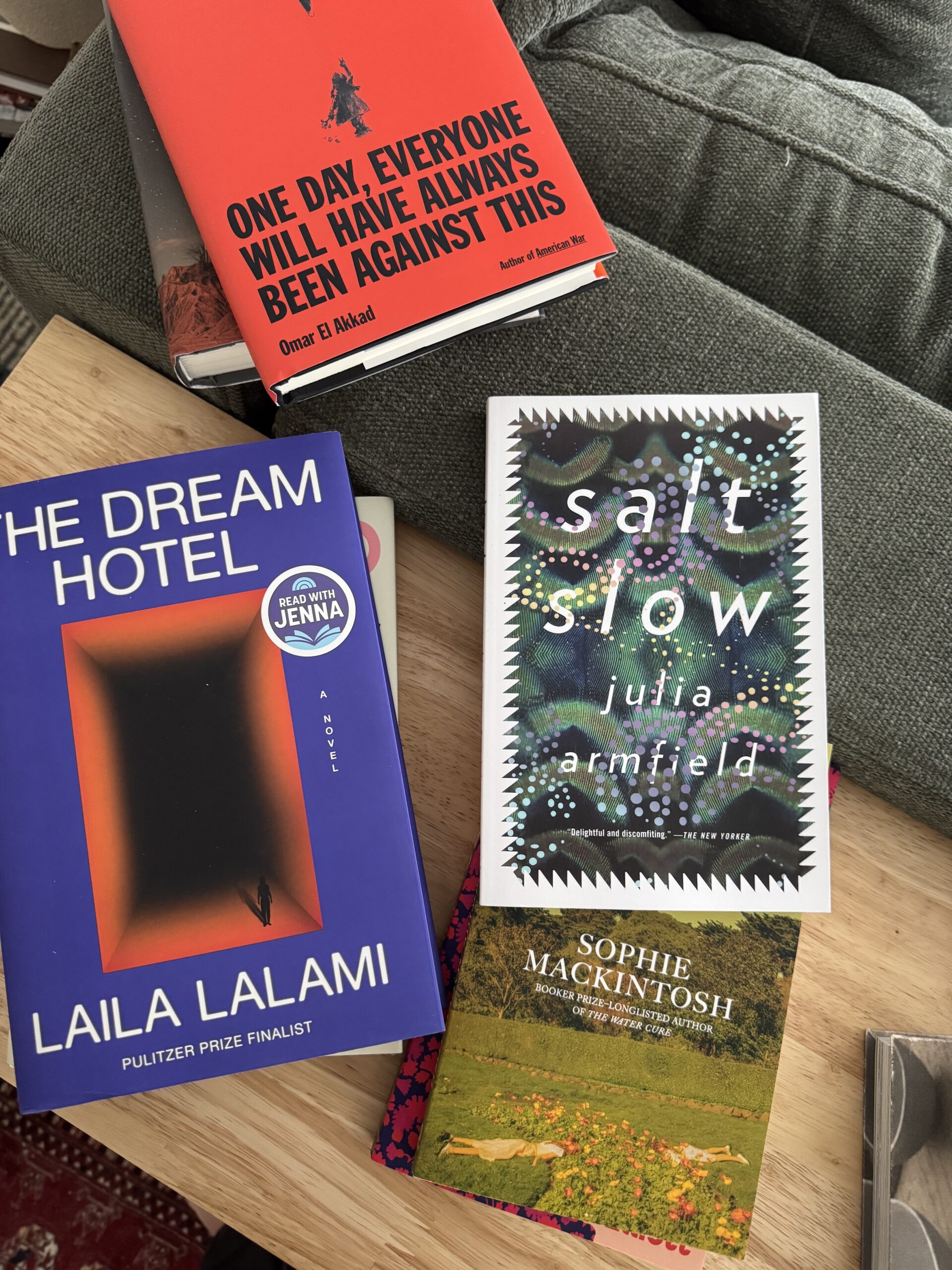 Four books—"The Dream Hotel," "Salt Slow," "One Day Everyone Will Have Always Been Against This," and "Cursed Bread"—are arranged on a wooden table near a green sofa cushion.