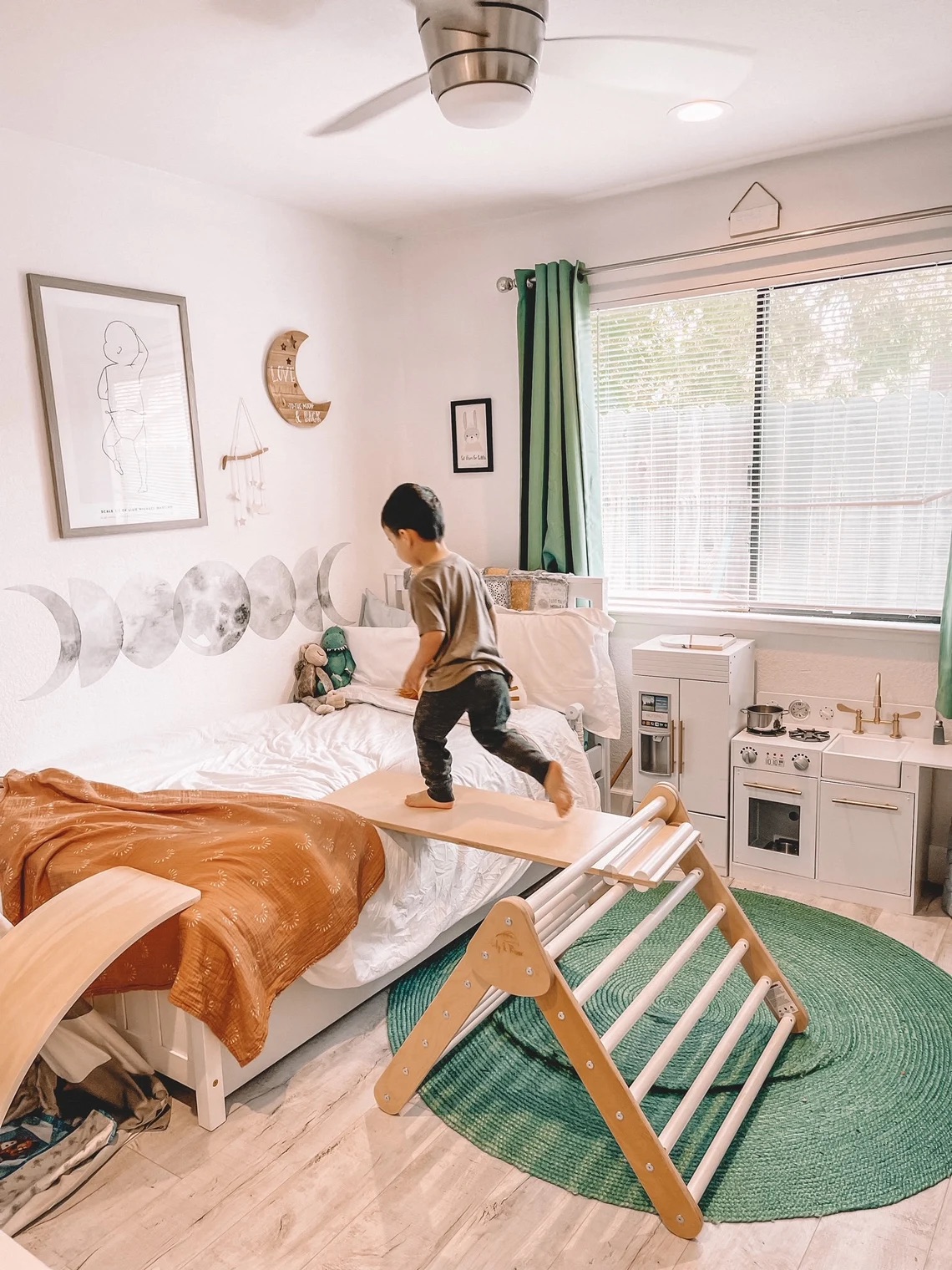 A child stands on a wooden climbing frame next to a bed in a bright, neatly decorated bedroom. The room has a green round rug, a toy kitchen set, and moon-themed decor on the walls.
