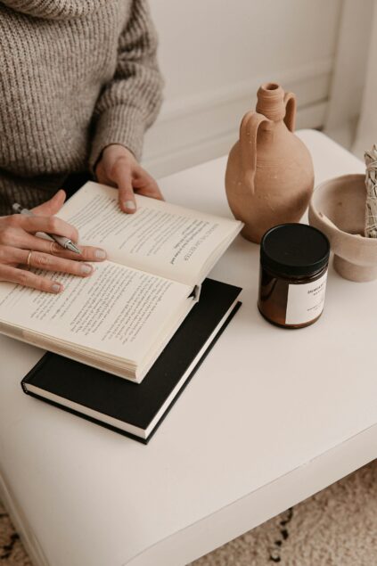 A person seated, reading a book with a table displaying a clay pot, a candle, and a notebook.