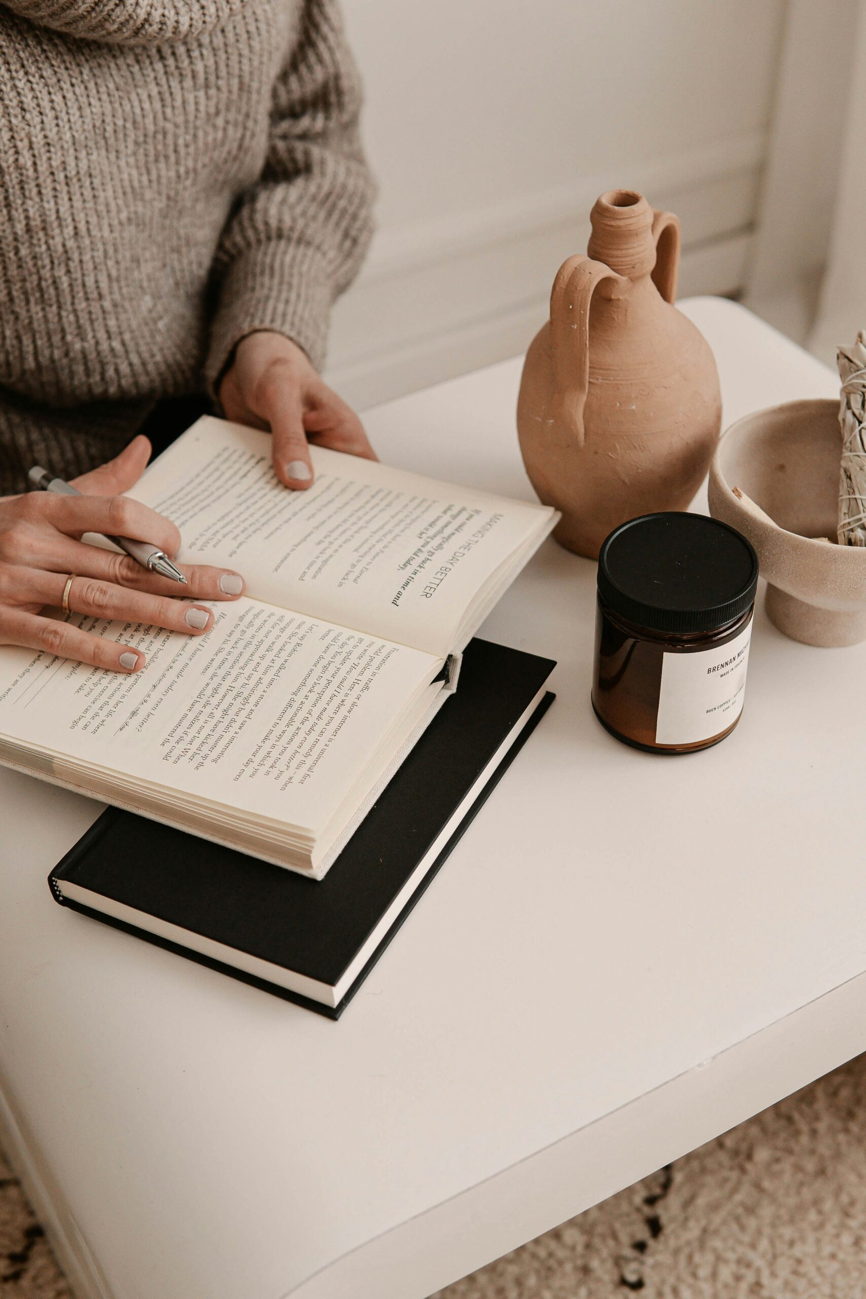 A person seated, reading a book with a table displaying a clay pot, a candle, and a notebook.