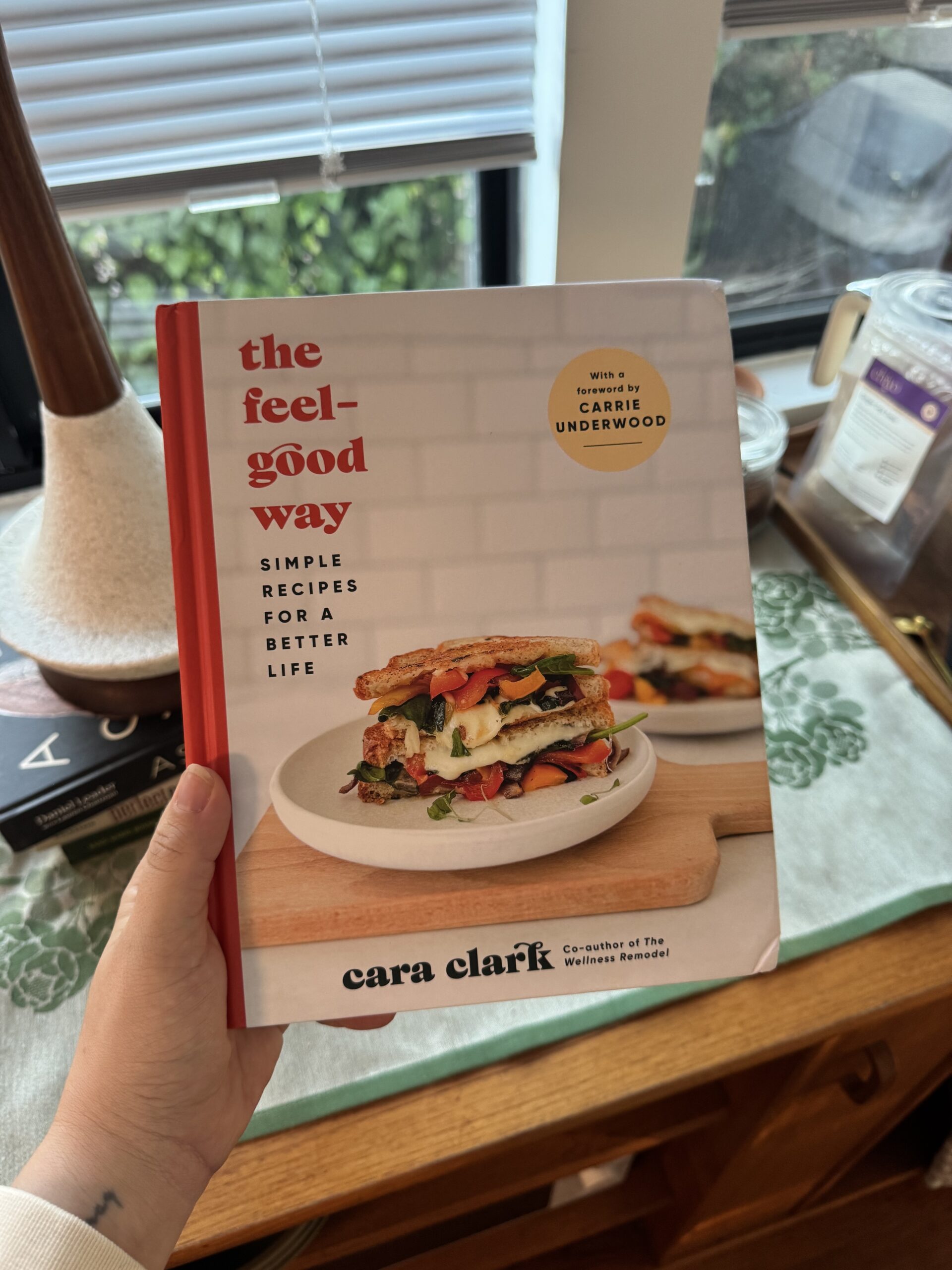 A hand holds the cookbook "The Feel-Good Way" by Cara Clark, featuring a sandwich on the cover and the subtitle "Simple Recipes for a Better Life.