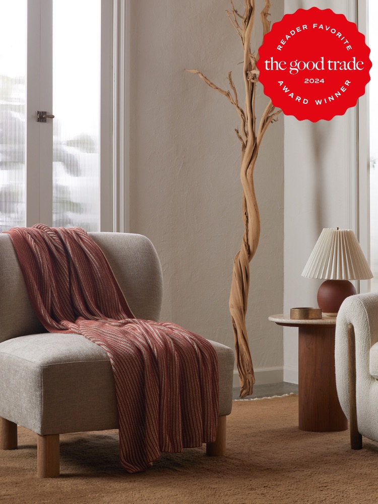 A cozy living room features a modern beige armchair draped with a red throw, next to a small wooden table with a lamp. A plant is visible in the background. A red award badge is in the top right corner.