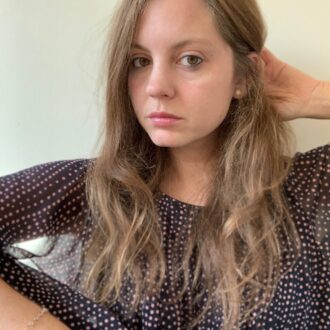 A woman with long, wavy brown hair wearing a black polka dot shirt looks at the camera while touching her hair. She has a neutral expression and is positioned against a plain background.