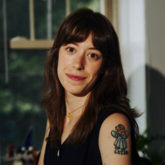A woman with long brown hair and bangs, wearing a sleeveless dark top, displays a tattoo of a cartoon character with the name "Luiza" on her upper arm. She is smiling, with a window in the background.