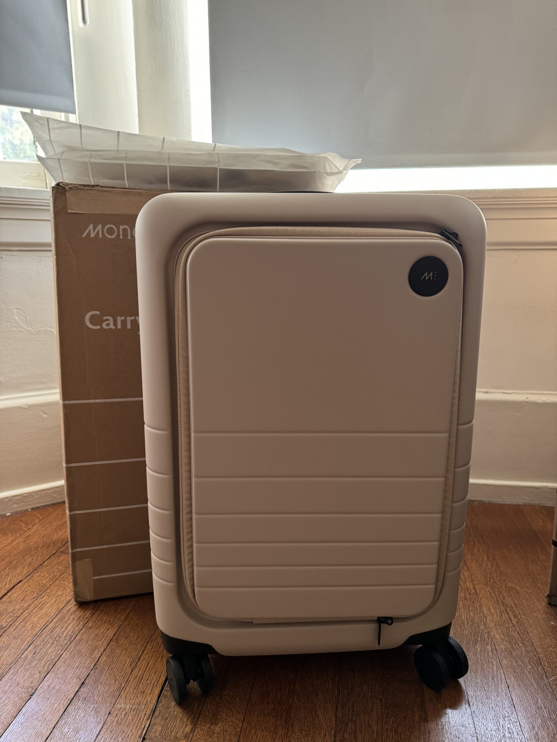A light pink hard-shell carry-on suitcase stands on a wood floor in front of a cardboard box near a window.