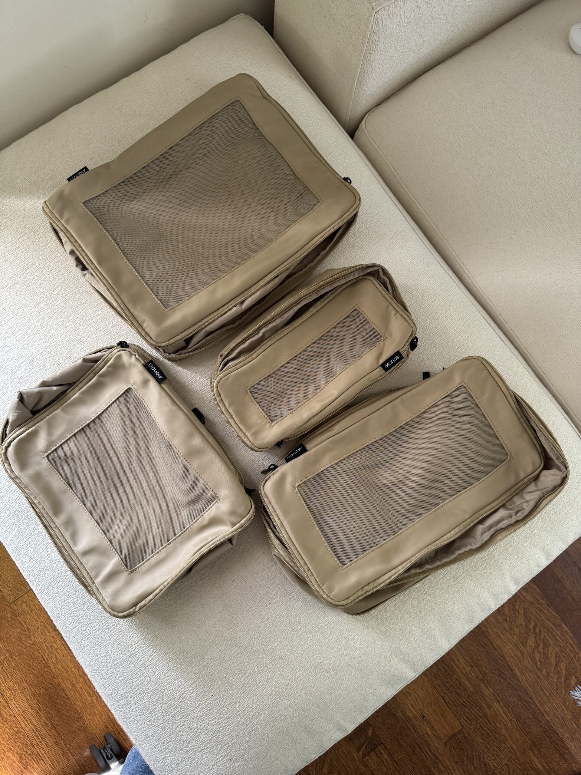 Four beige packing cubes of varying sizes are arranged on a white cushioned surface, with a wooden floor visible in the background.