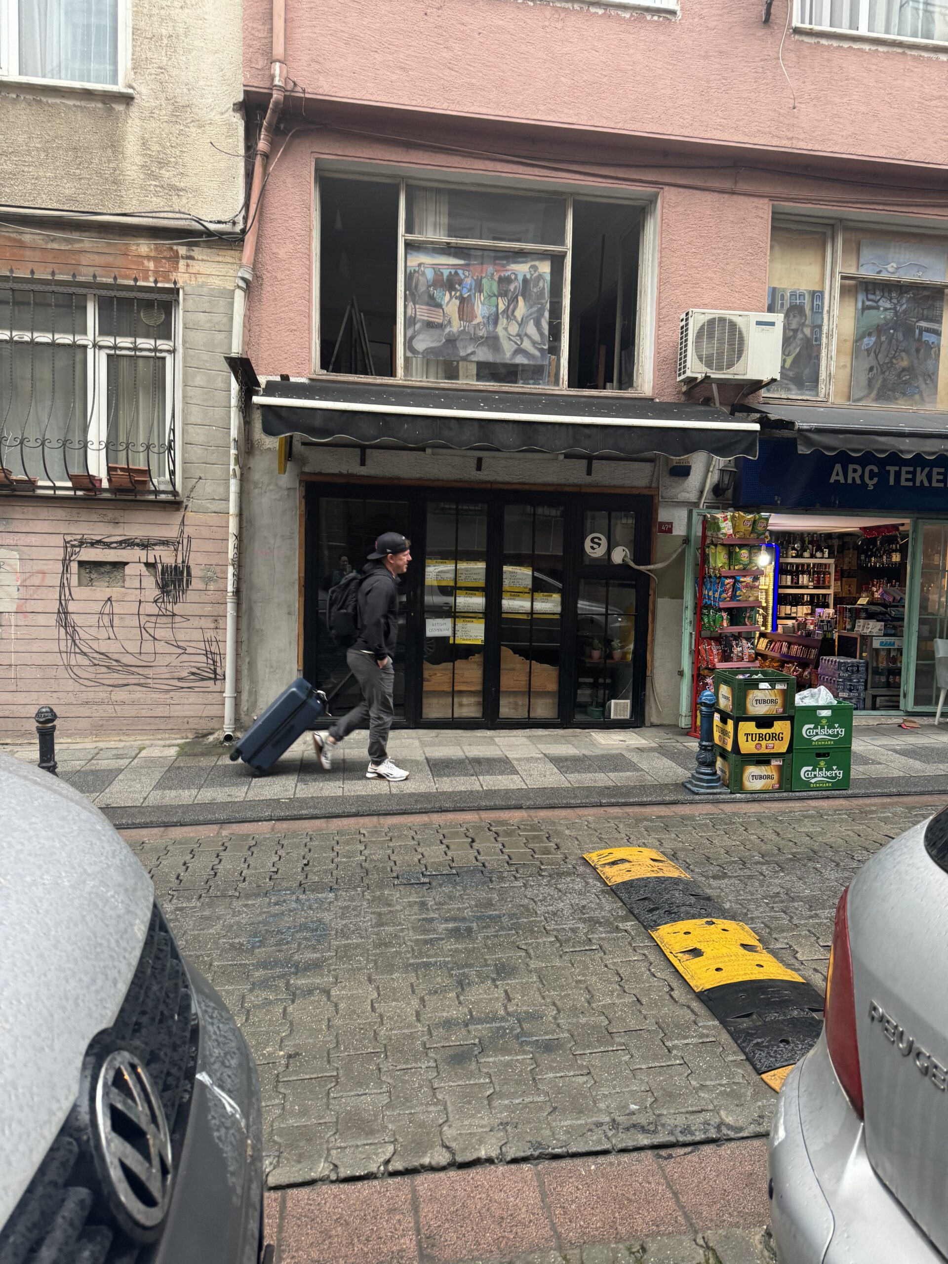 Person walking down a street with a rolling suitcase in front of a small storefront, next to a shop with beverages on display outside.