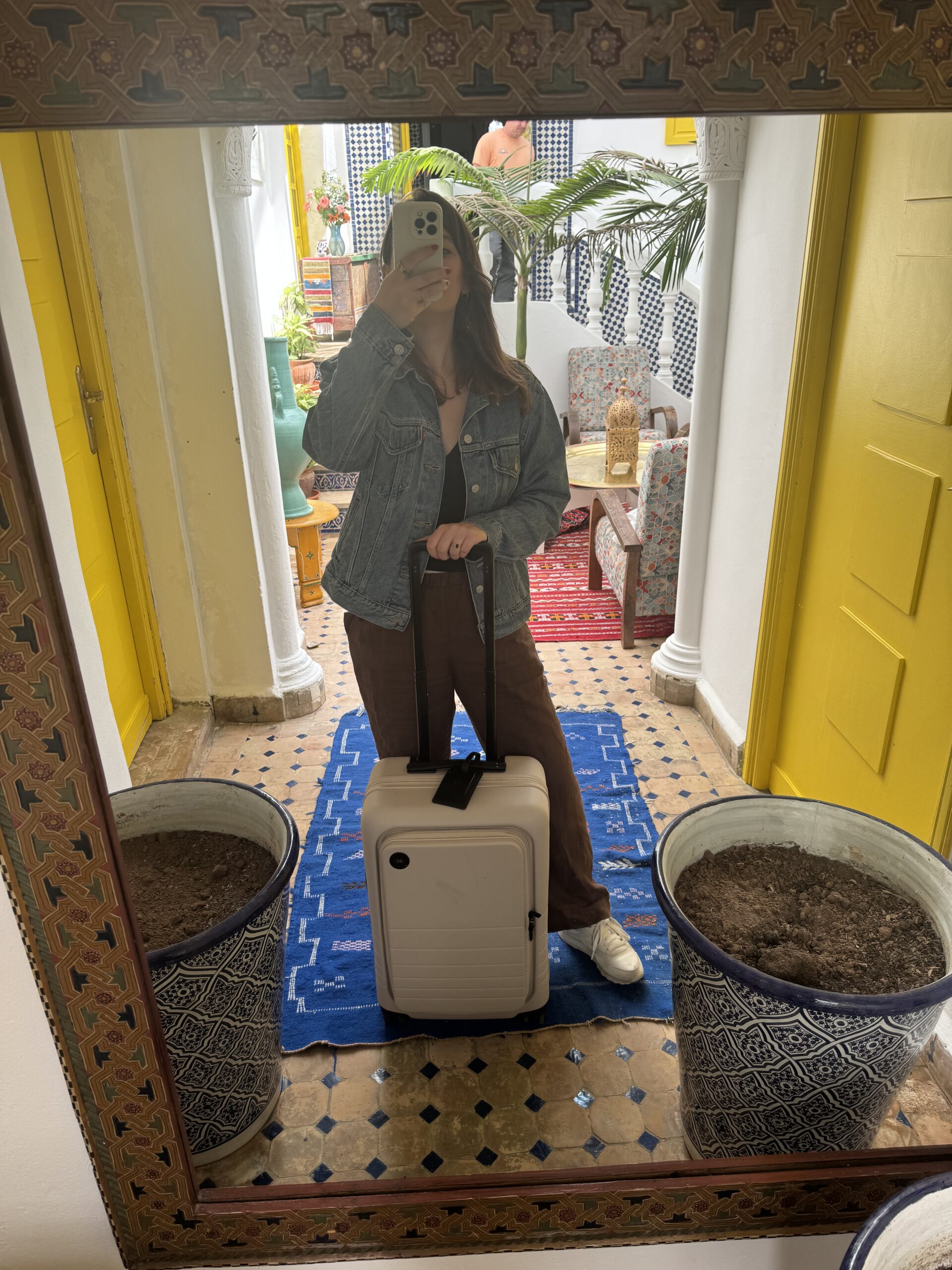 A person in a denim jacket and brown pants takes a mirror selfie while holding a suitcase handle in a room with yellow doors and blue, white, and brown patterned decor.