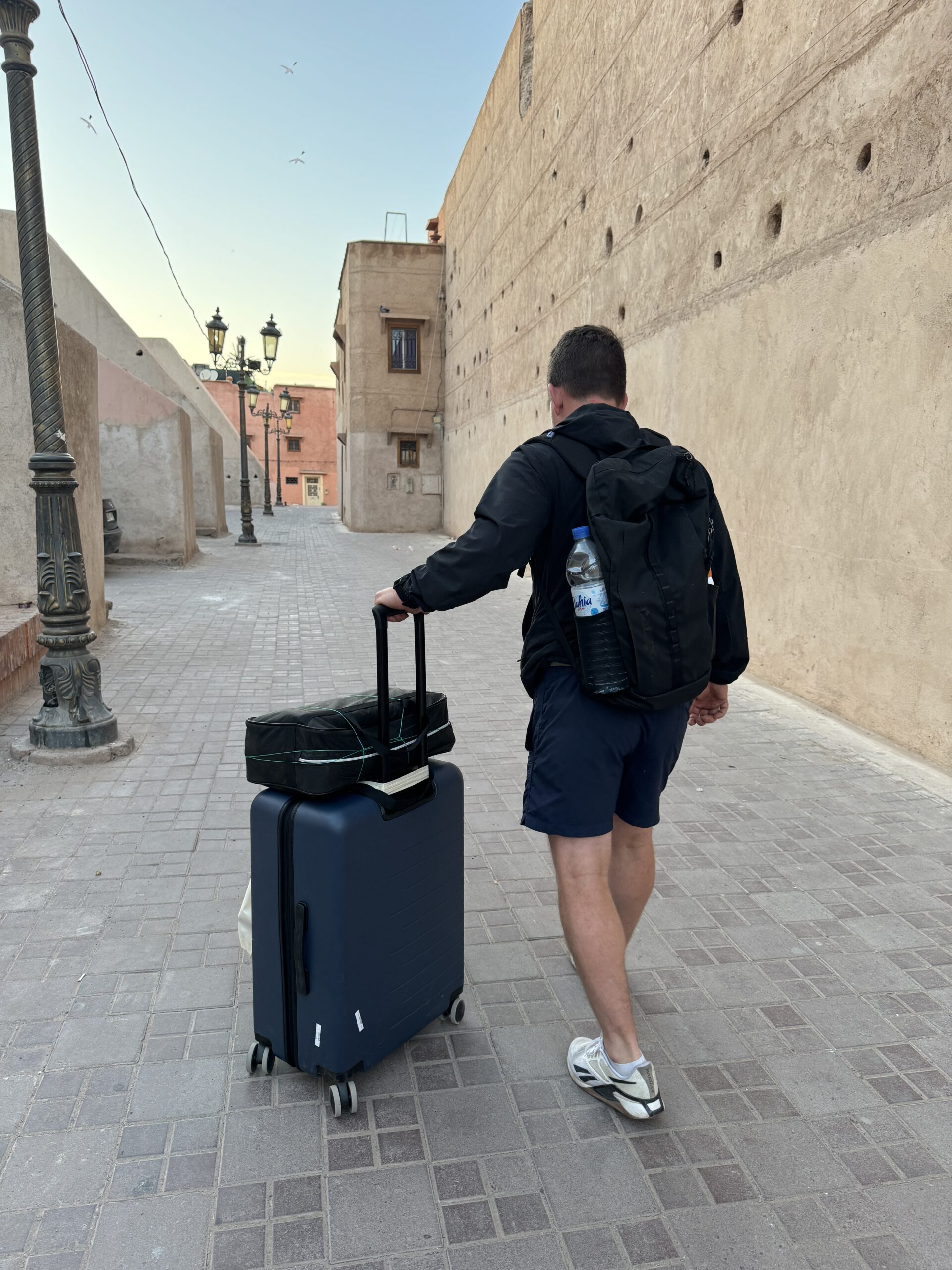 A person in casual attire walks down a narrow, empty street with a suitcase and a backpack, approaching a historic-looking building with light poles lining the walkway.