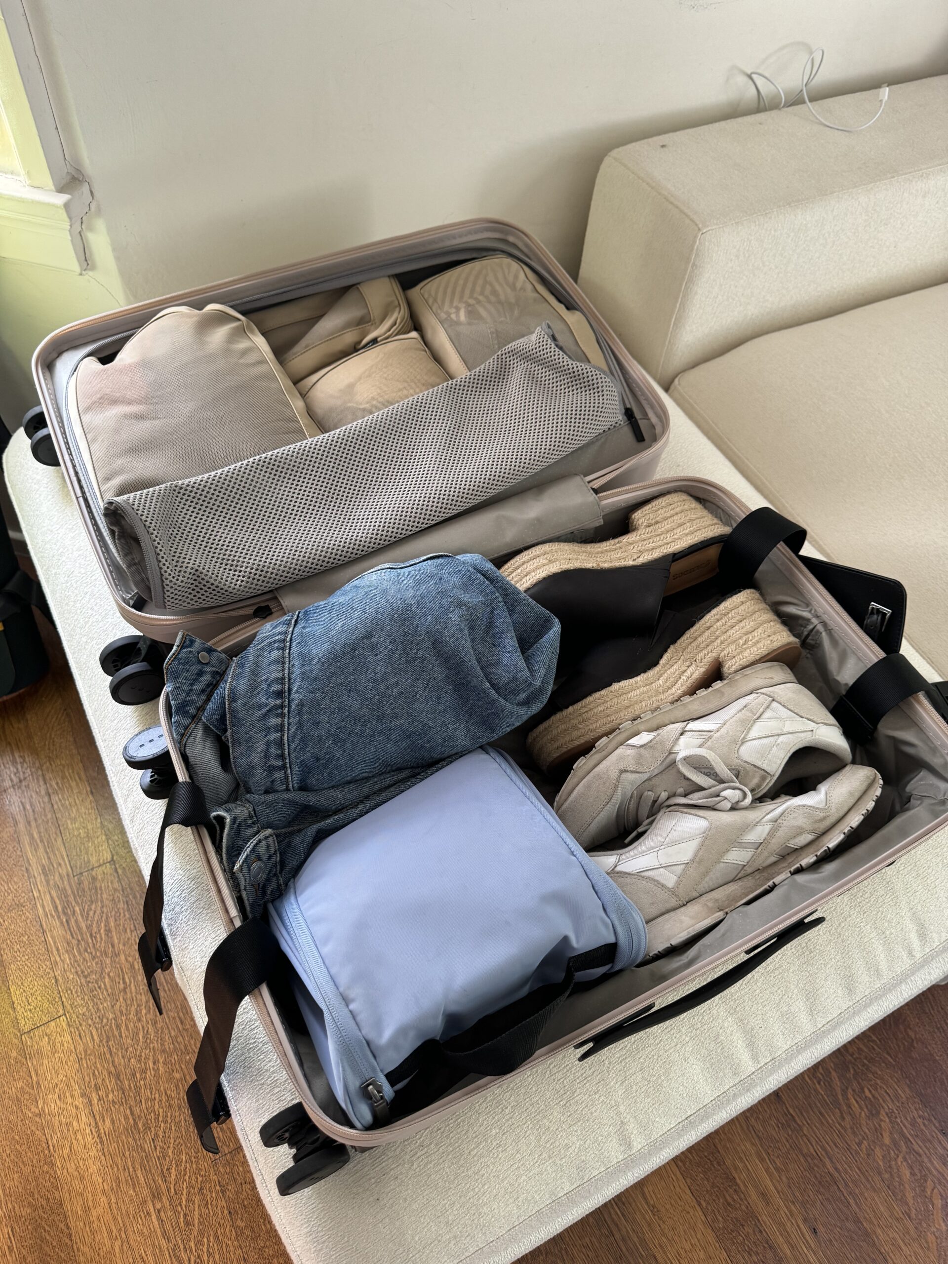 An open suitcase is packed with shoes, a backpack, a small blue bag, denim shorts, and various folded clothes, placed on a white surface indoors.