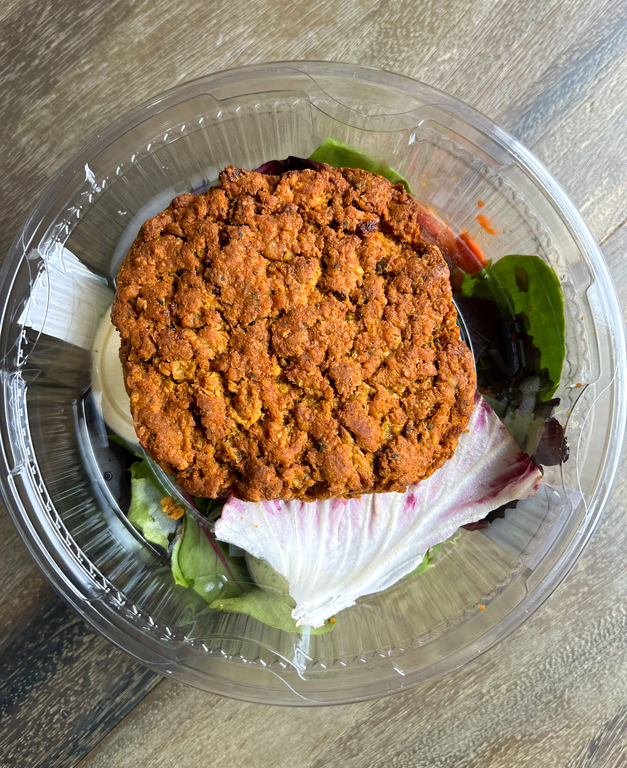 A plastic container with a salad topped with a large falafel patty, mixed greens, and a leaf of radicchio on a wooden surface.