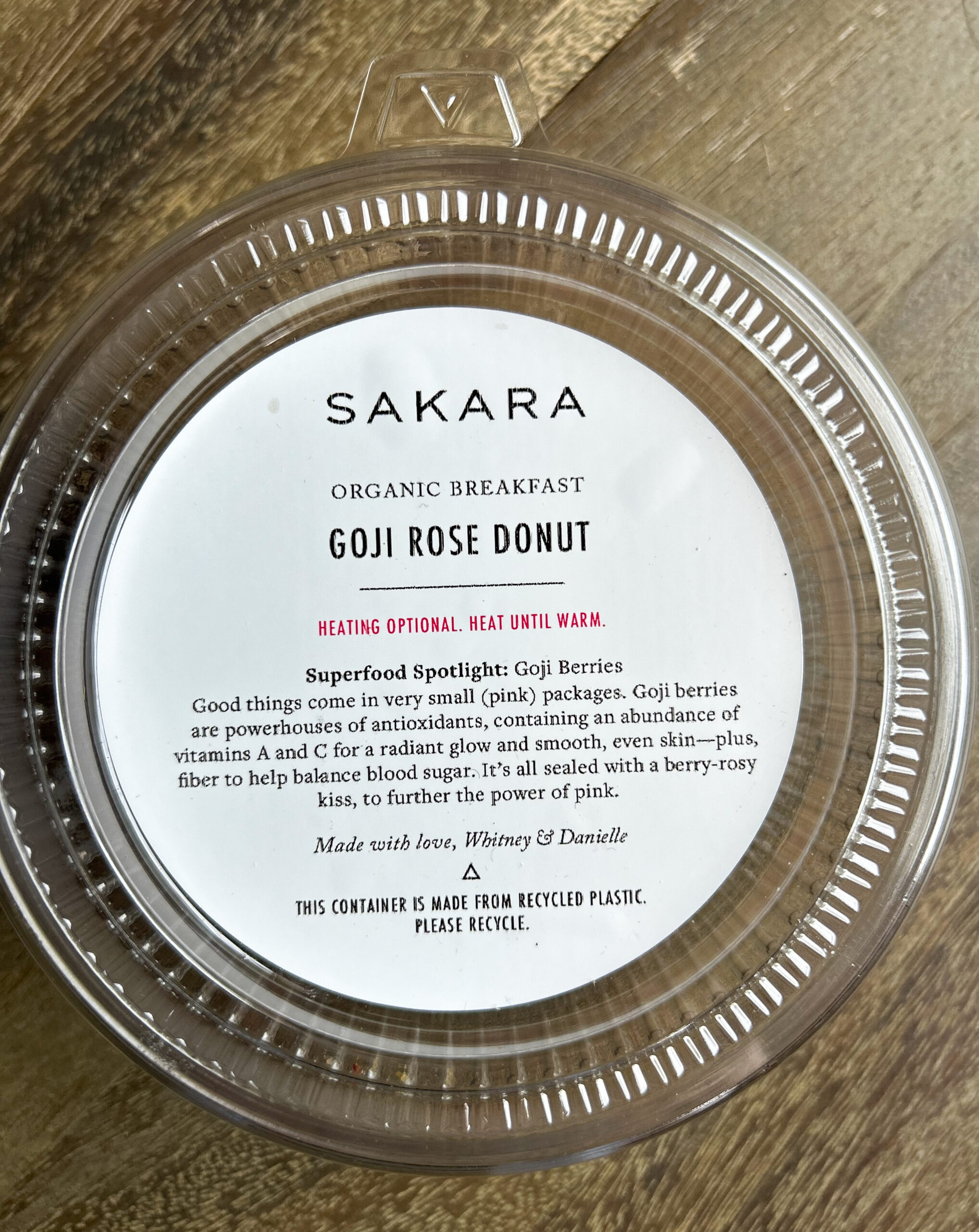 Round plastic container with a label that reads "Sakara Organic Breakfast Goji Rose Donut" and includes heating instructions and details about goji berries. The container is recyclable.