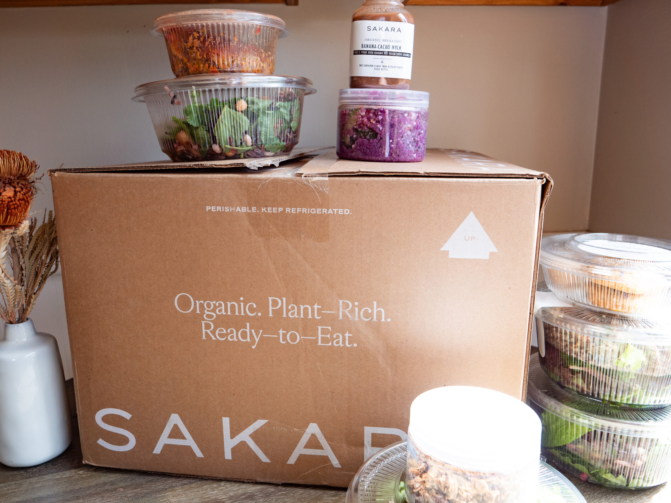 A large cardboard box labeled "SAKARA" with smaller containers of food on and around it. The box reads "Organic. Plant-Rich. Ready-to-Eat." The containers feature various prepared meals.